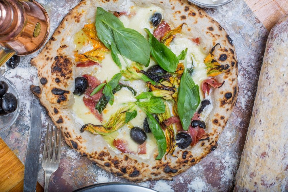 Wood-fired pizza topped with basil, olives, and vegetables on a flour-dusted table