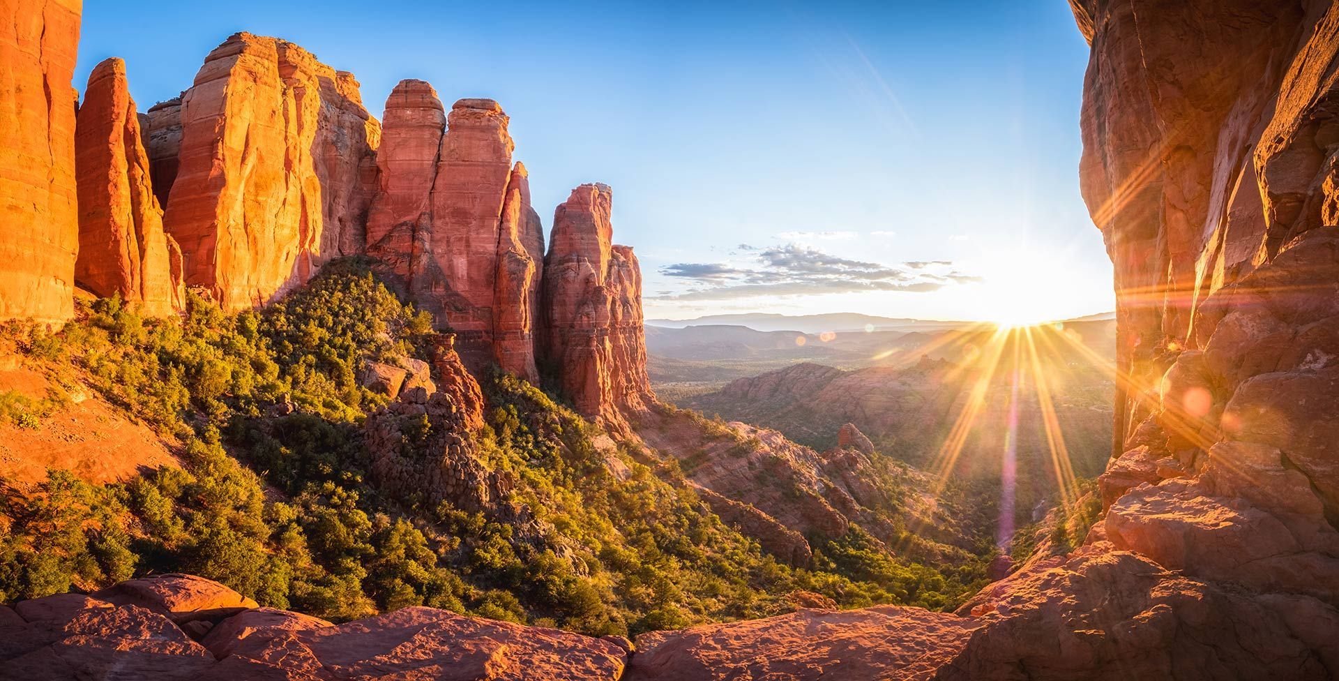 Sunrise over red sandstone cliffs and a green canyon, with sun rays streaming through a rocky arch