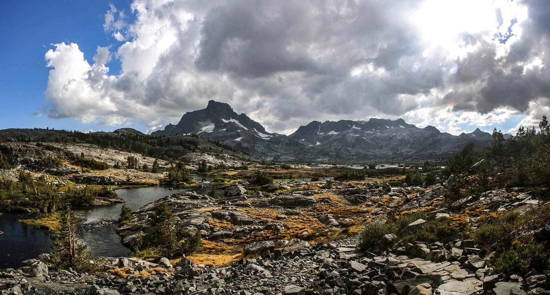Rocky mountain landscape with a river, cloudy sky, and scattered yellow wildflowers