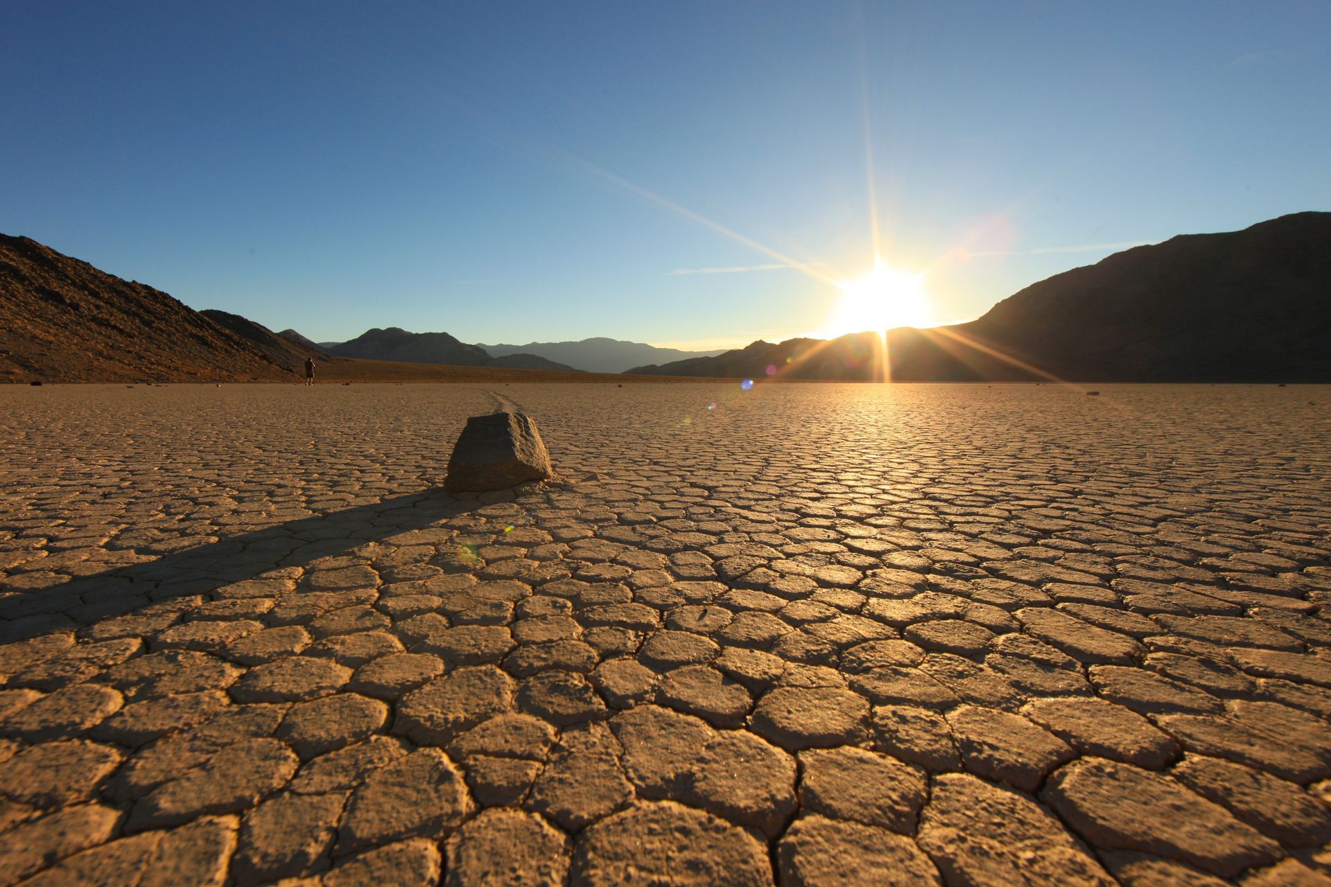 Sunset over cracked desert الأرض, with mountains on both sides and a small rock in the foreground