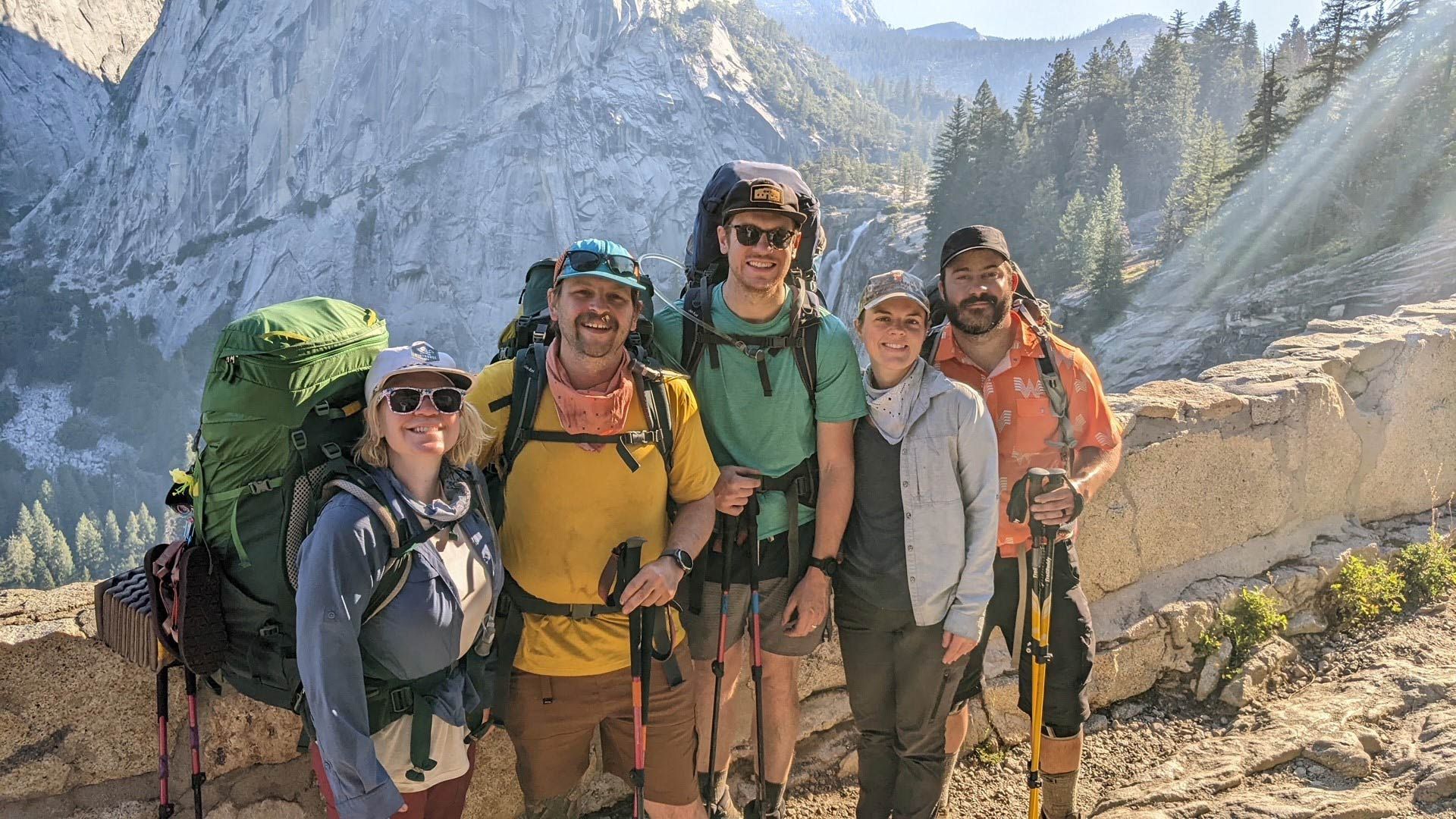 Group of hikers with backpacks posing on a trail with a mountain backdrop.