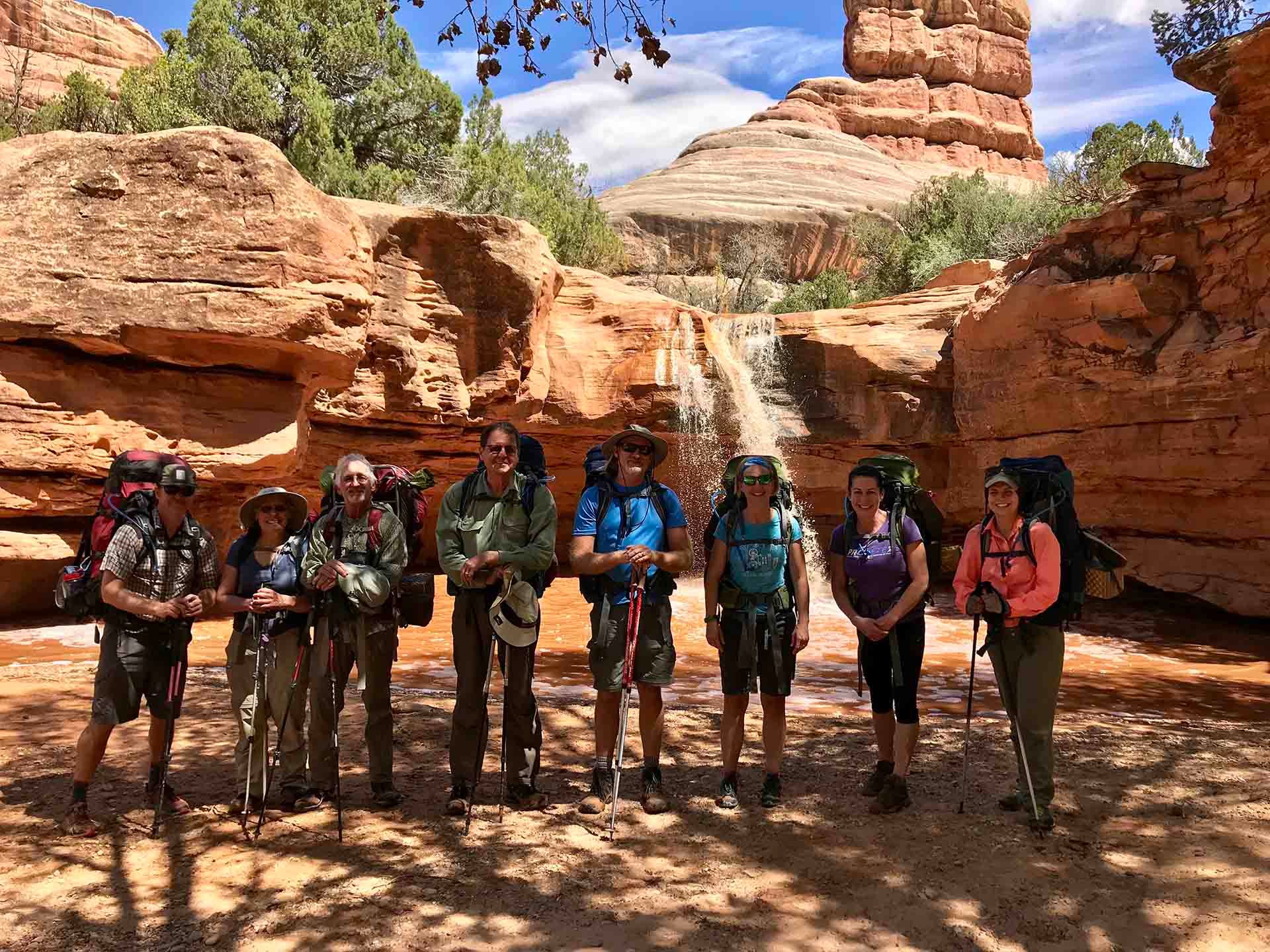 Group of hikers in canyon, backpacks, trekking poles. Waterfall in background, red rock formations.