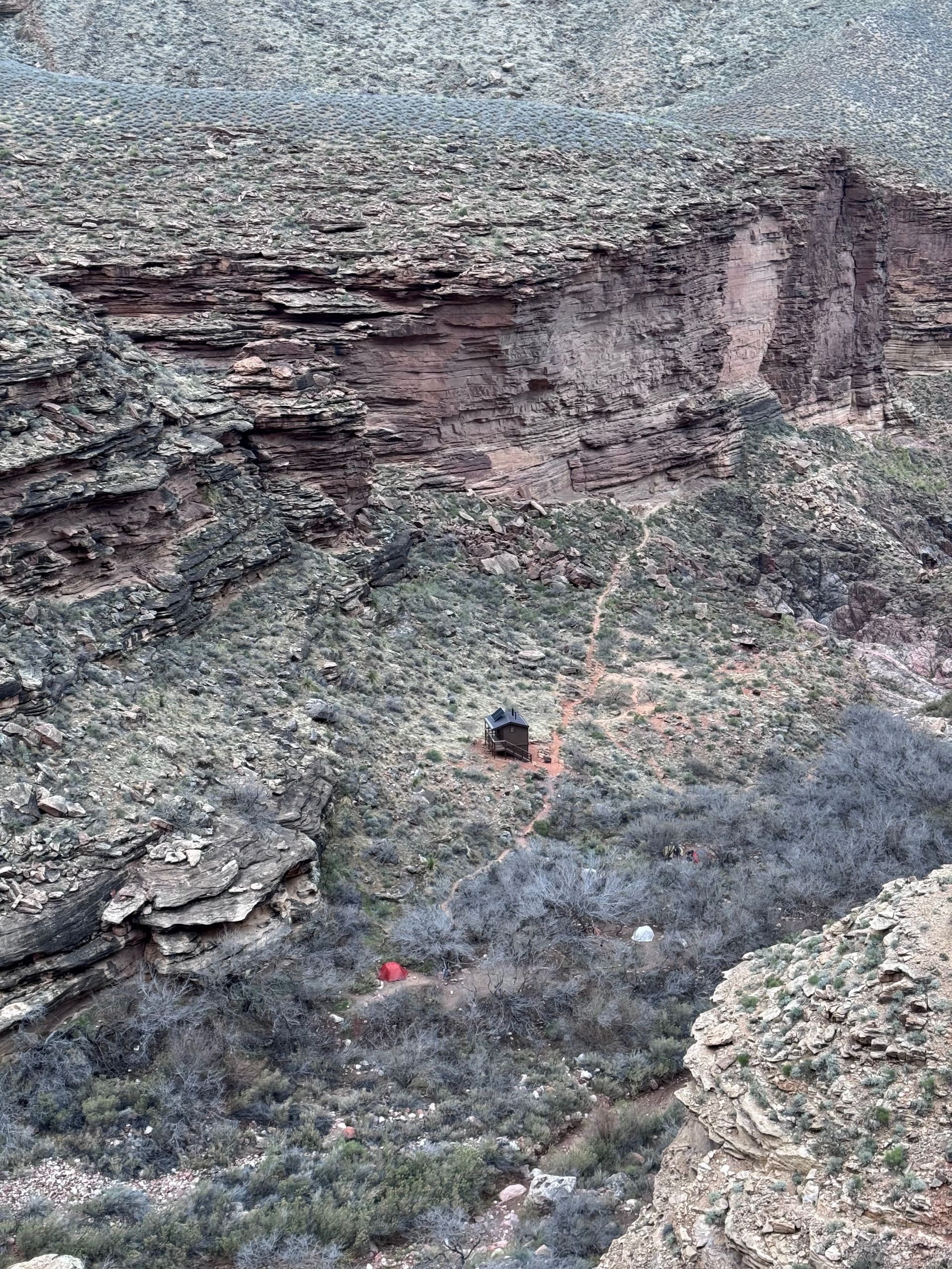 Rocky landscape with a person and a red object near the bottom. Tan and gray cliffs in the background.