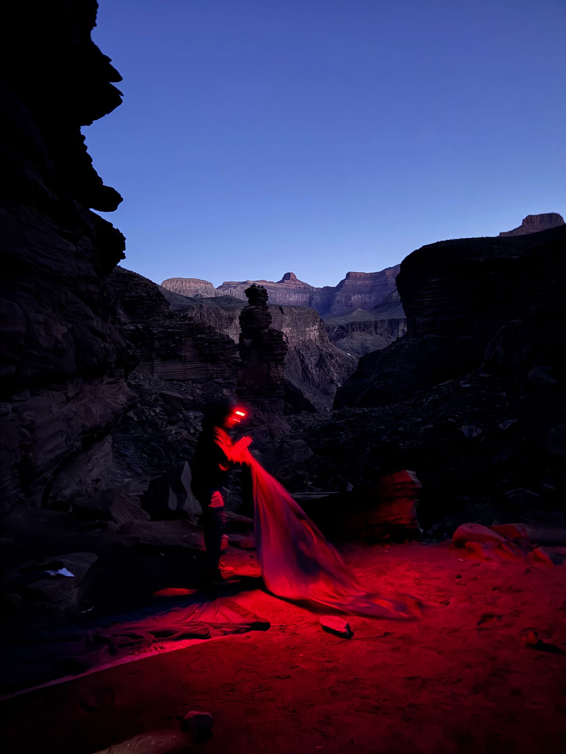 Person with headlamp in a canyon at dusk, illuminating red light. Rocky landscape under a blue sky.