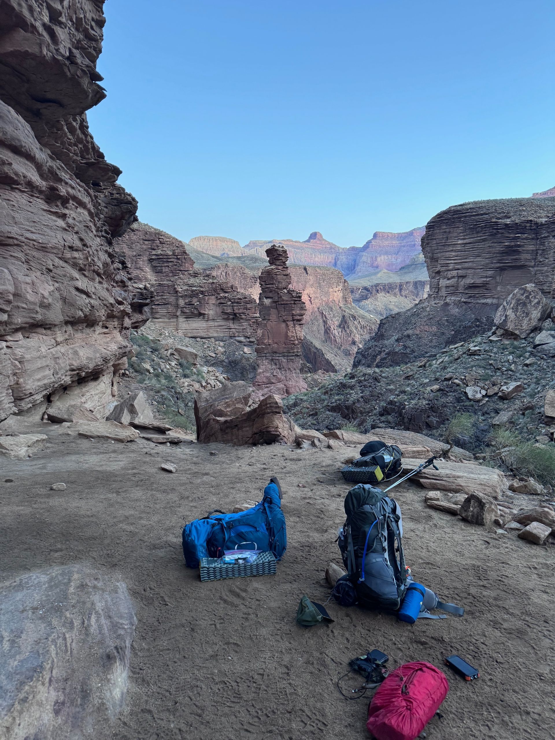 Backpacks and gear on a sandy ledge overlooking a canyon with a rock spire, under a blue sky.