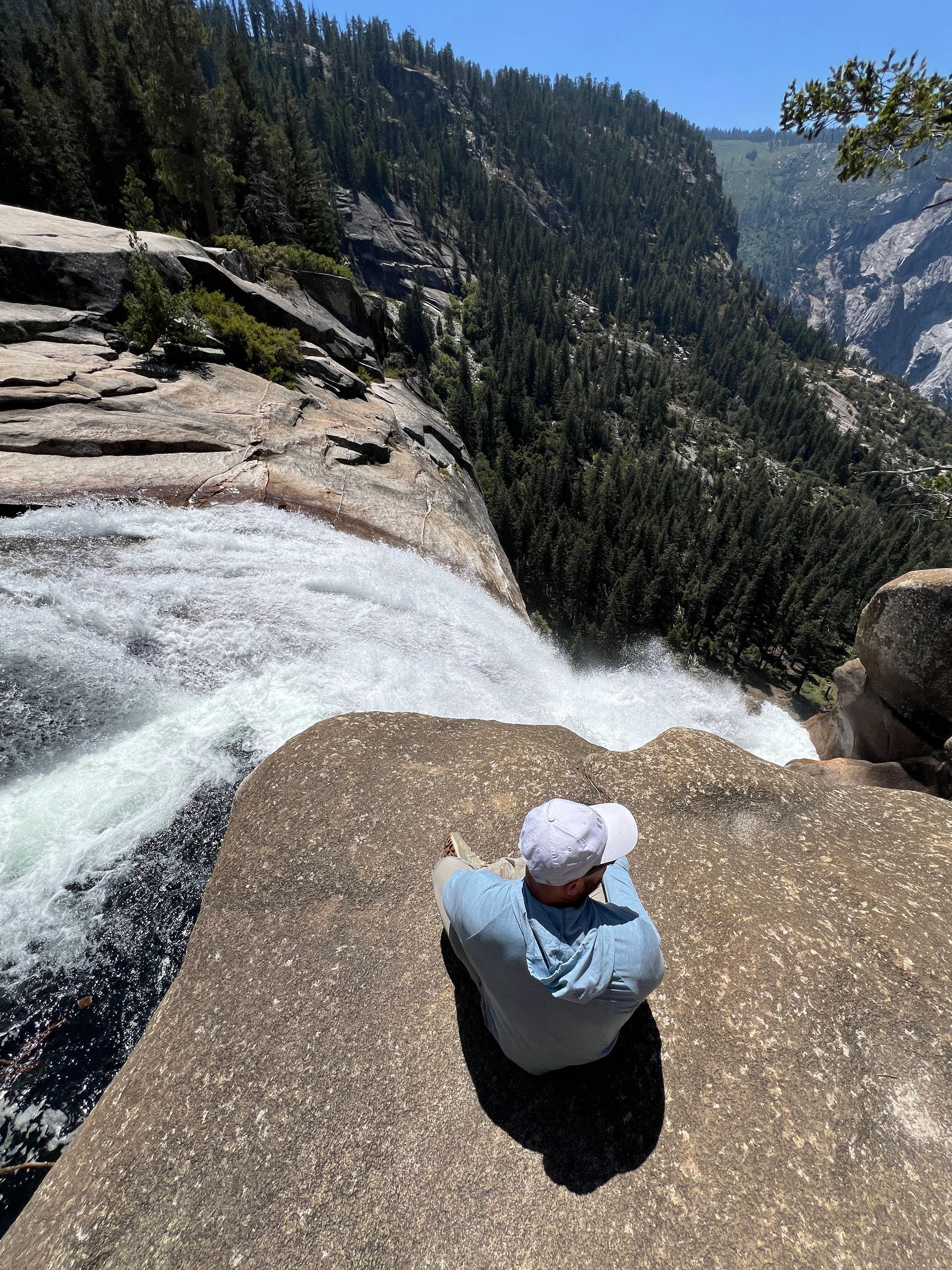 Person sits at edge of waterfall, looking down. Rocky terrain, blue sky, trees in background.