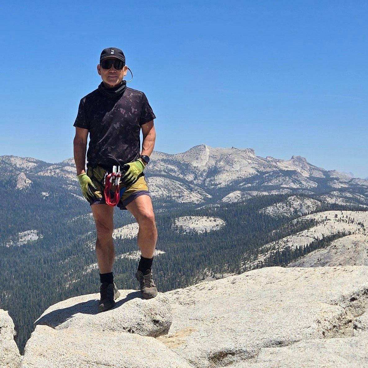 Man stands on a rock summit, mountain backdrop. Blue sky, green trees. Wearing sunglasses, black shirt, shorts, gloves.