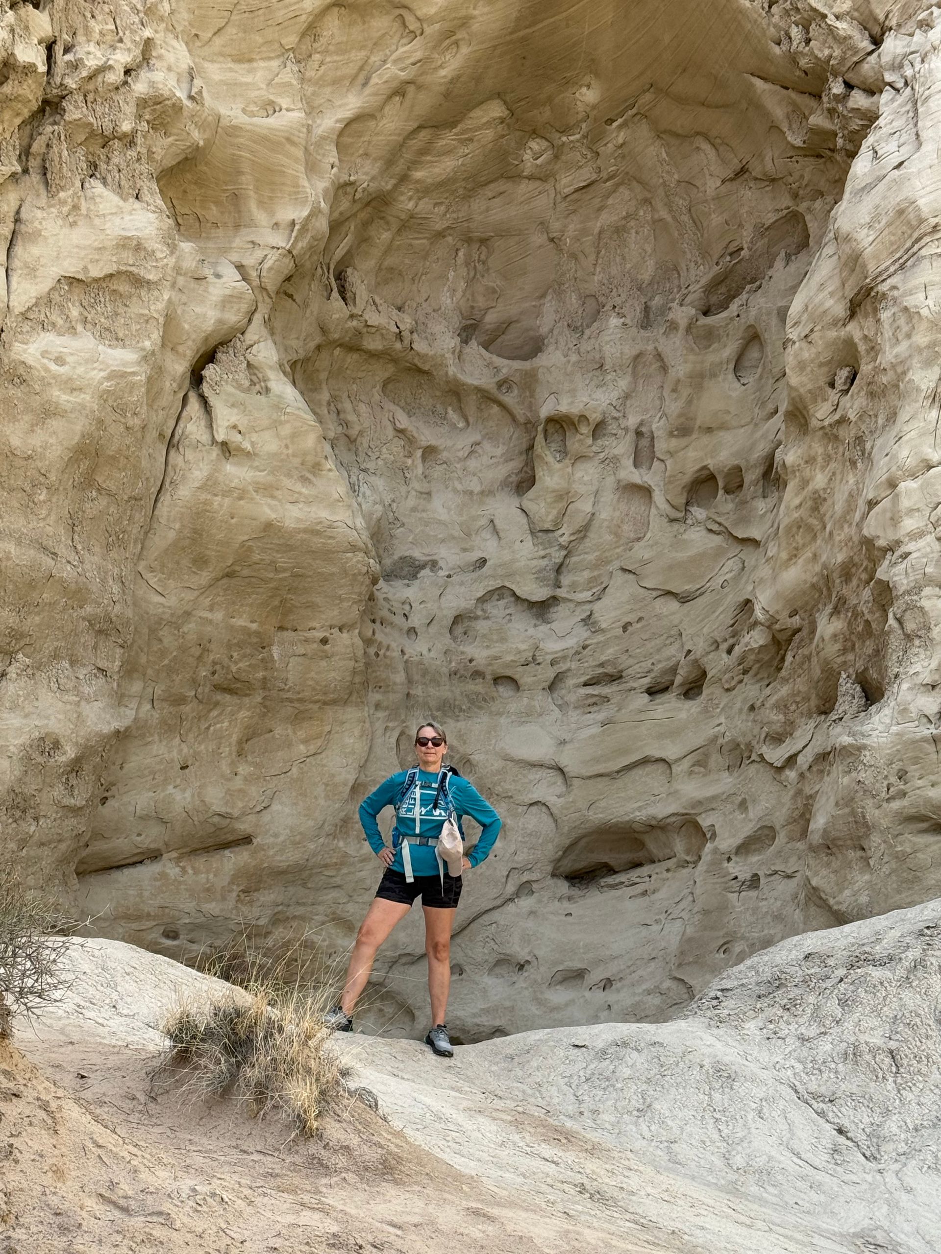 Woman standing in front of a tan rock wall formation, wearing a teal jacket and black shorts.