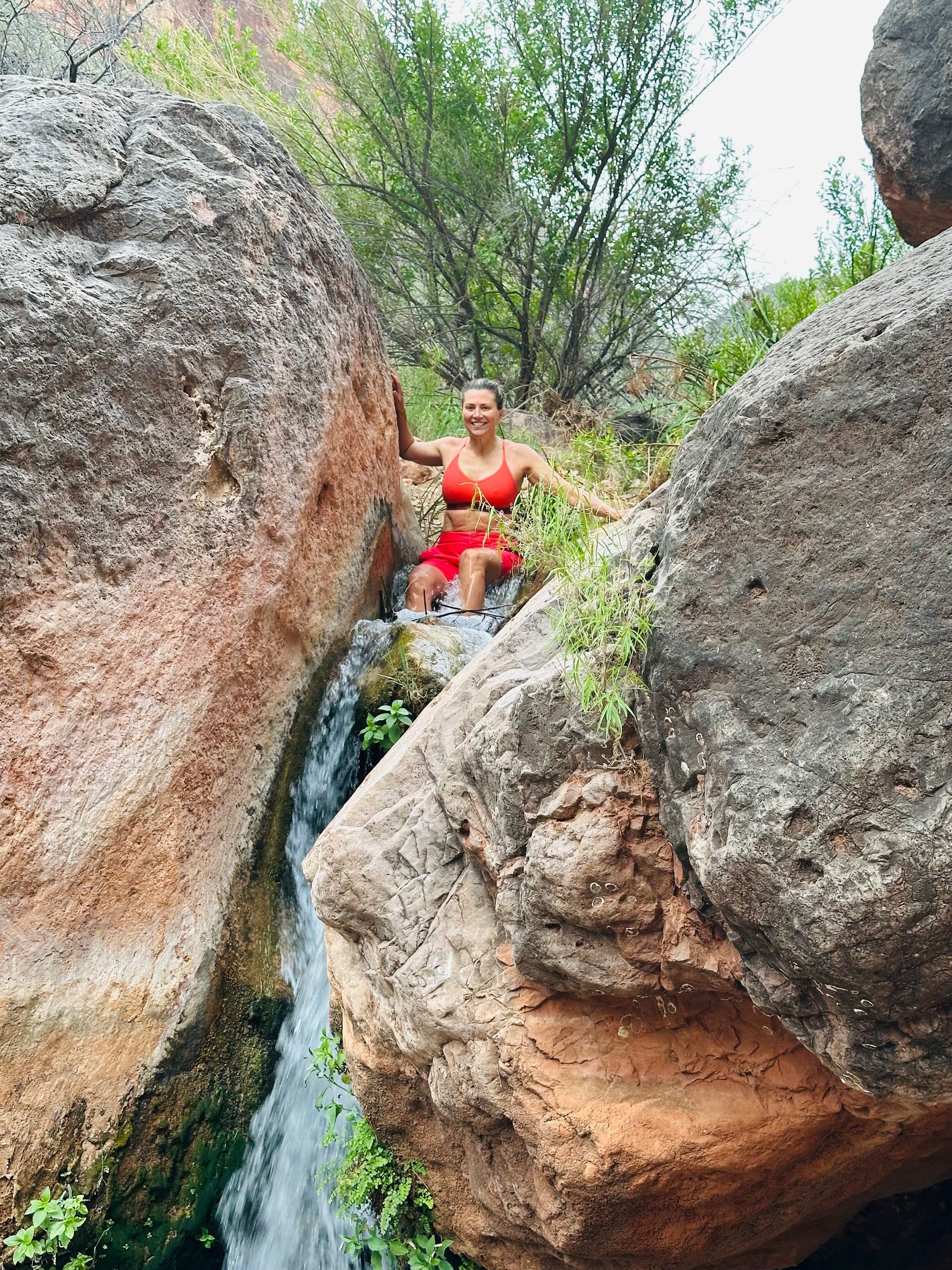 Woman in red outfit near a waterfall cascading between large rocks in a natural setting.