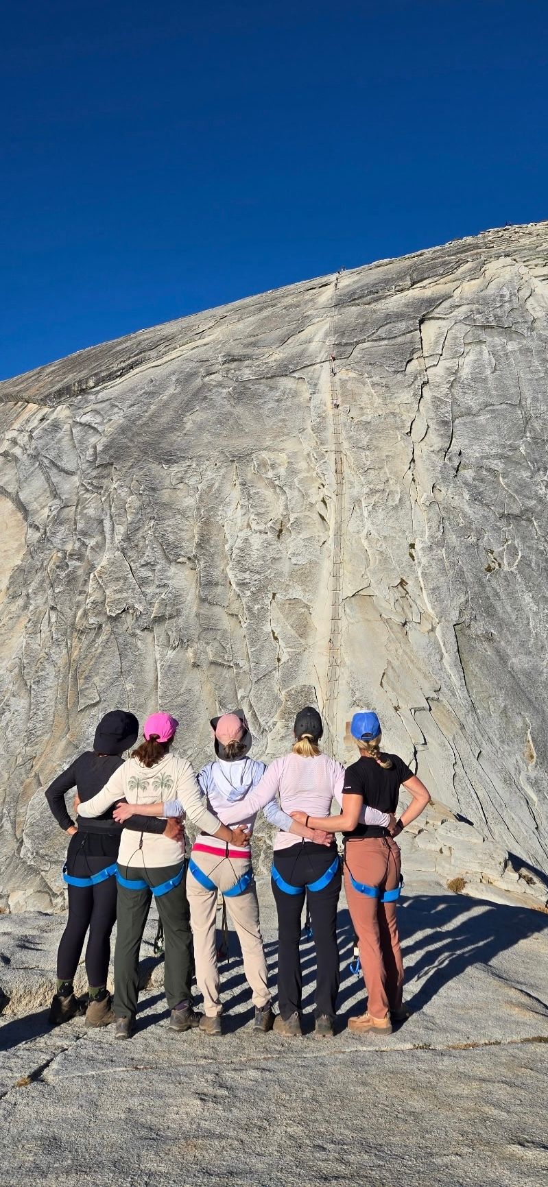 Five people in climbing gear stand arm-in-arm in front of a rock face under a clear blue sky.