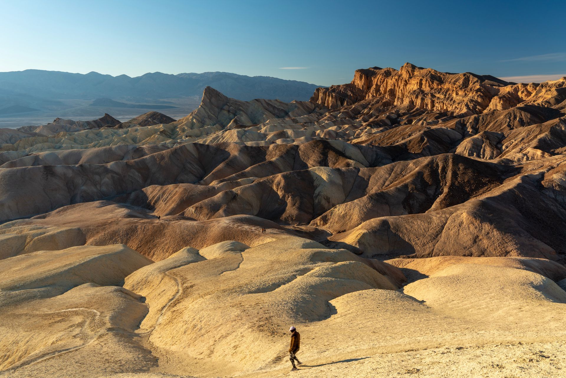 Man walking in a desert landscape with colorful, eroded formations under a blue sky.
