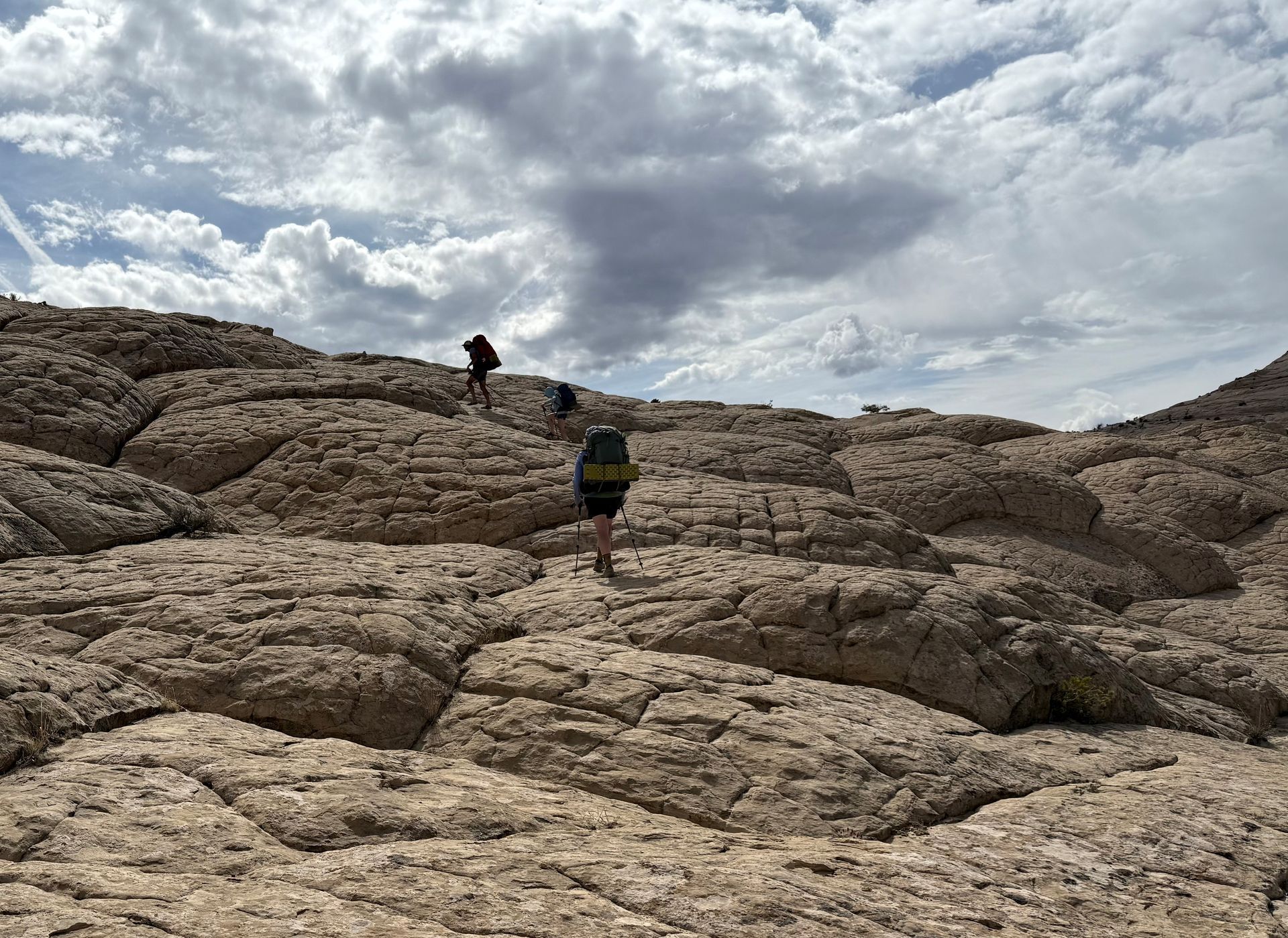 Hikers with backpacks ascend a rocky, textured slope under a cloudy sky.