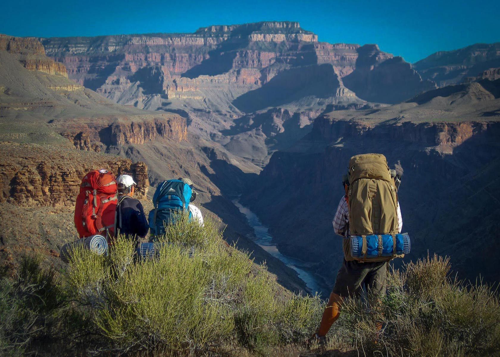Three hikers with large backpacks overlook a canyon under a clear blue sky.