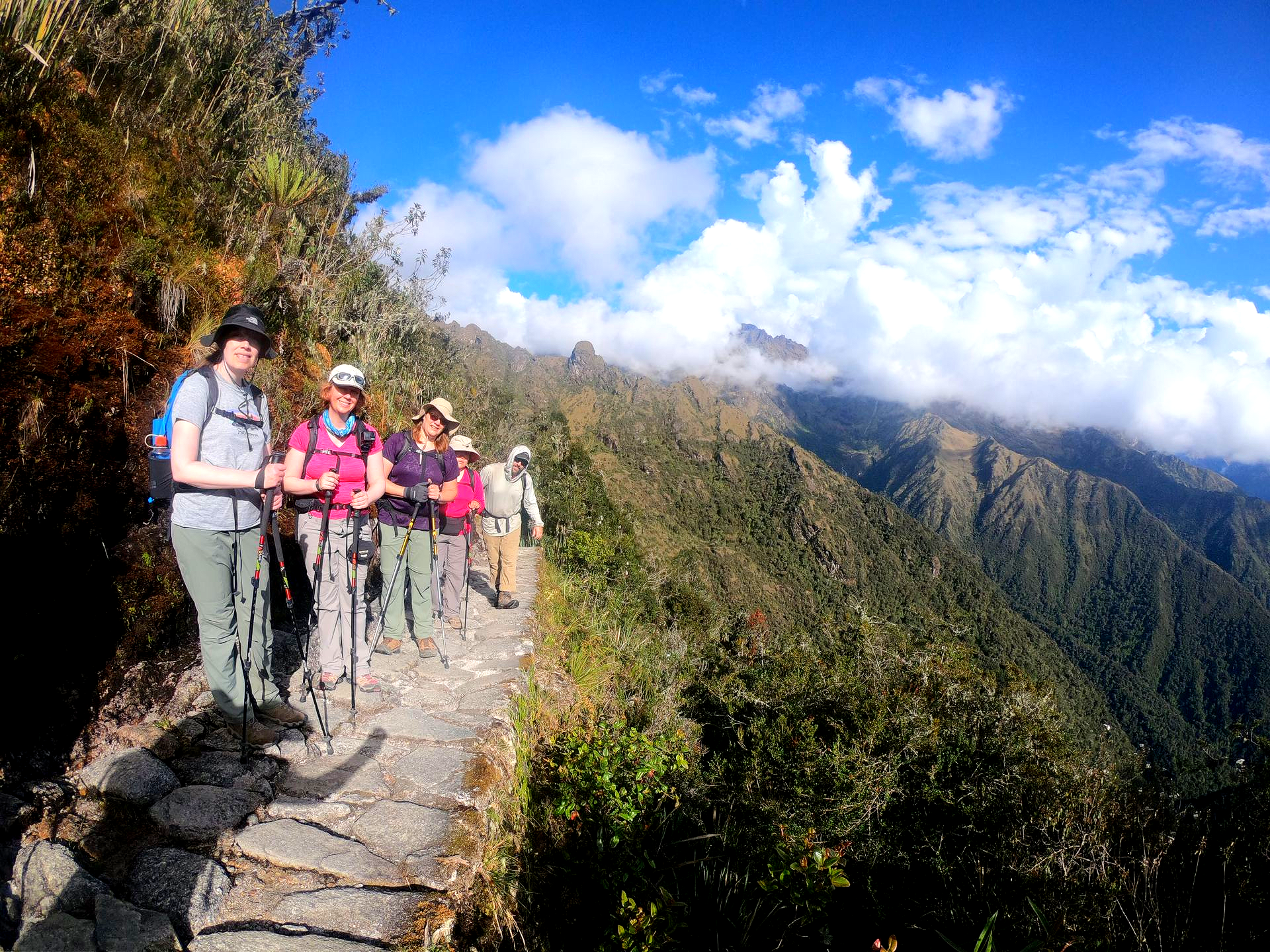 Hikers on a narrow mountain trail above a valley, with blue sky and clouds overhead.