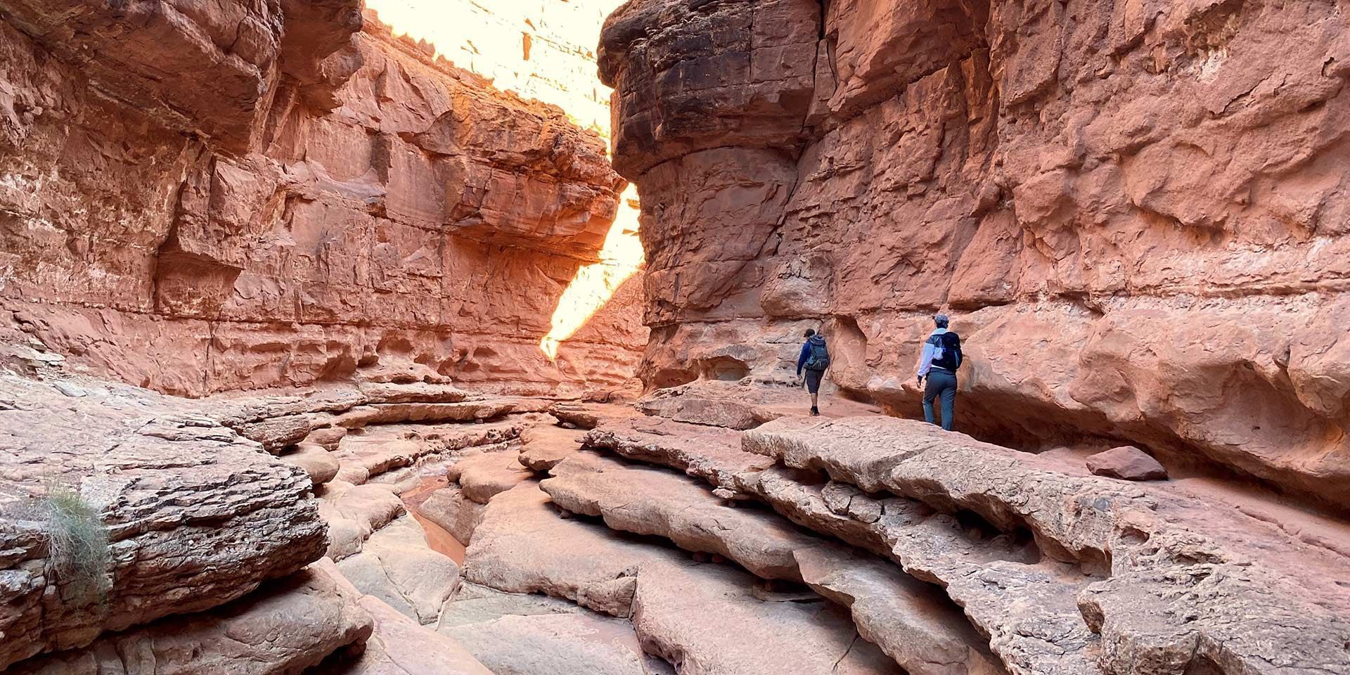 Hikers in a red rock canyon with layered sandstone and a bright opening above.