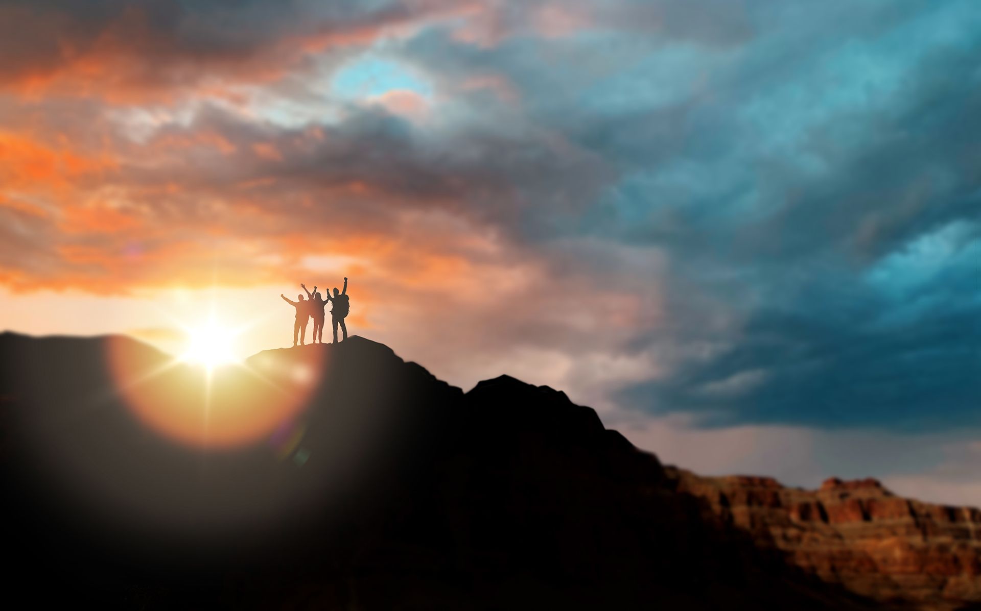 Silhouetted people on mountaintop, arms raised in celebration, sun setting behind them. Orange and blue sky.