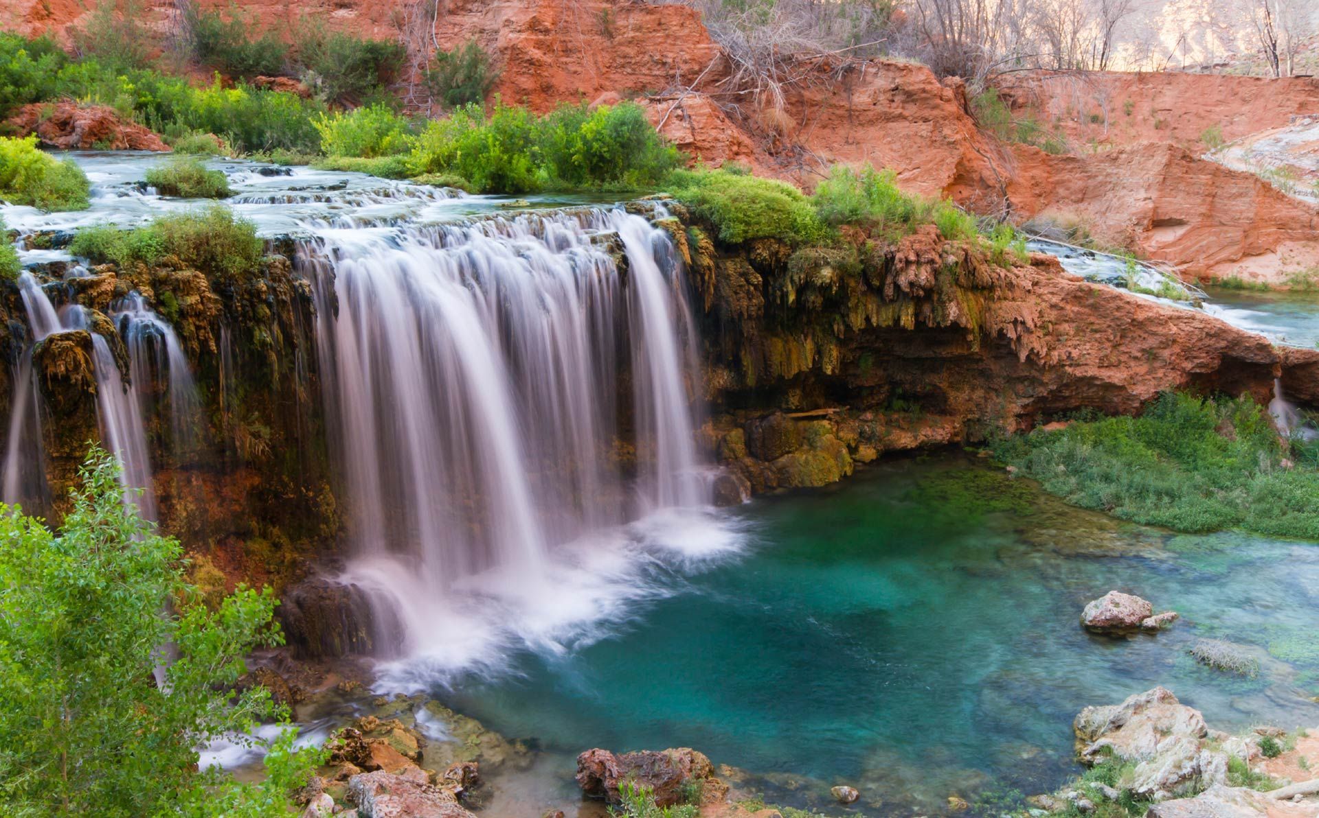 Small waterfall spilling into a turquoise pool beside red rock cliffs and green vegetation