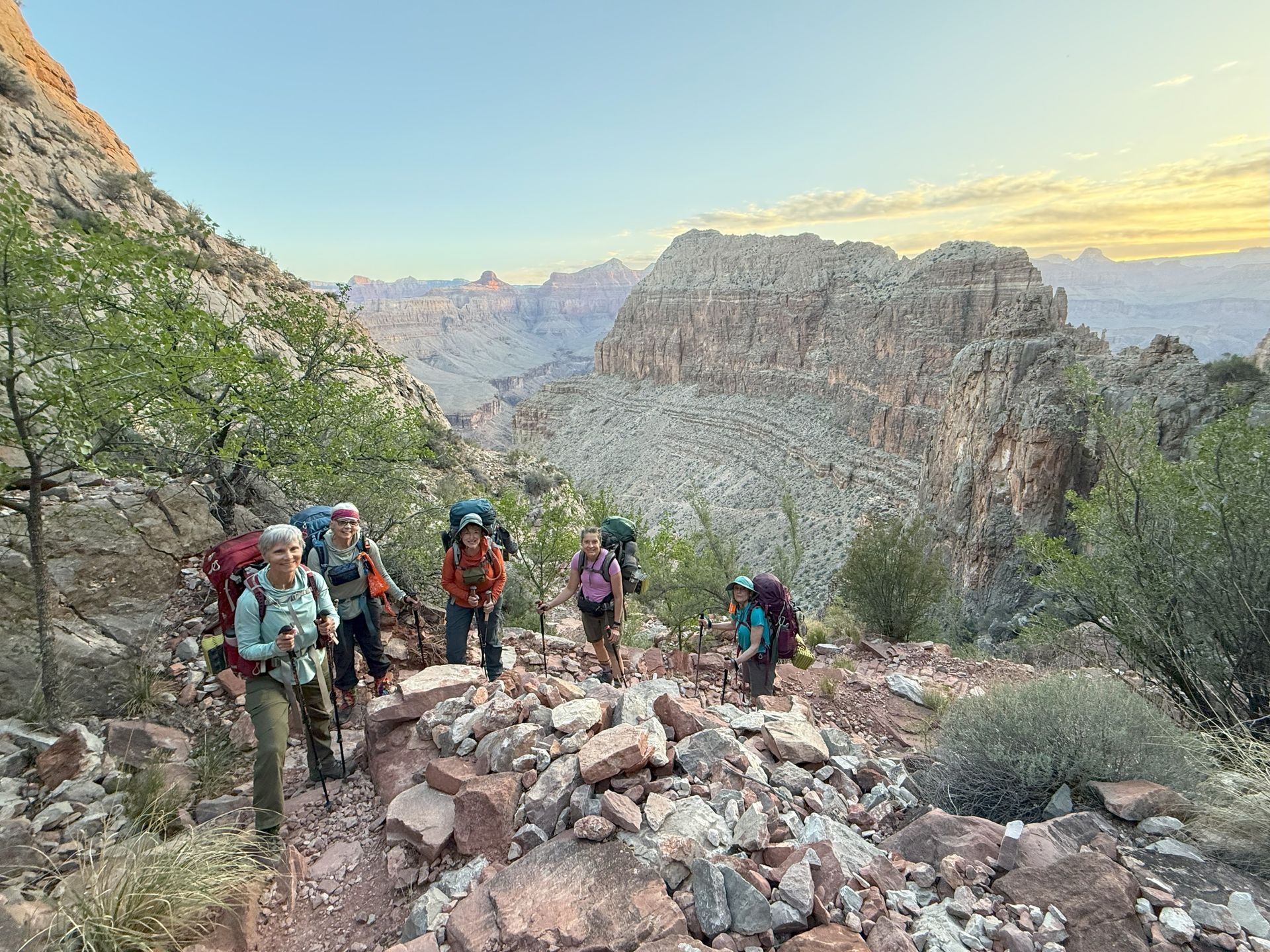 Hikers with backpacks on a rocky mountain trail at sunrise overlooking a canyon.