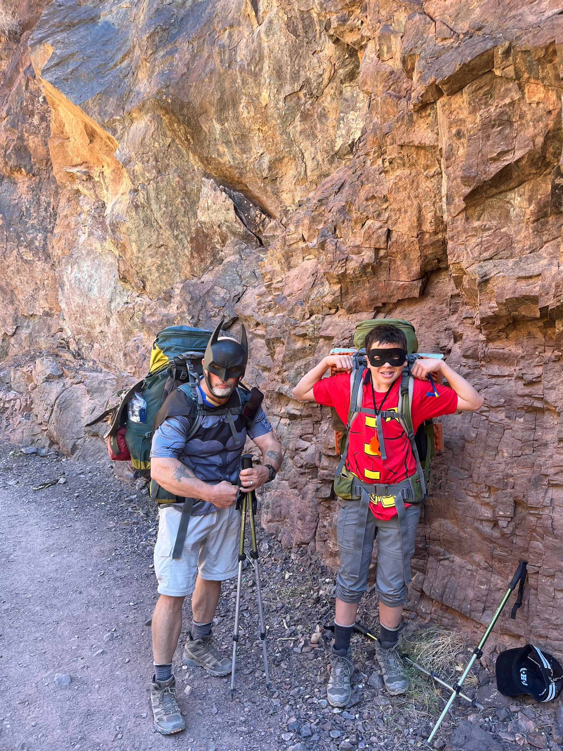 Two men with backpacks are standing next to each other in front of a rock wall.