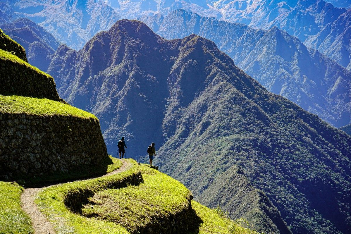 Two figures standing on a grassy mountain path, overlooking a range of green mountains under a blue sky.