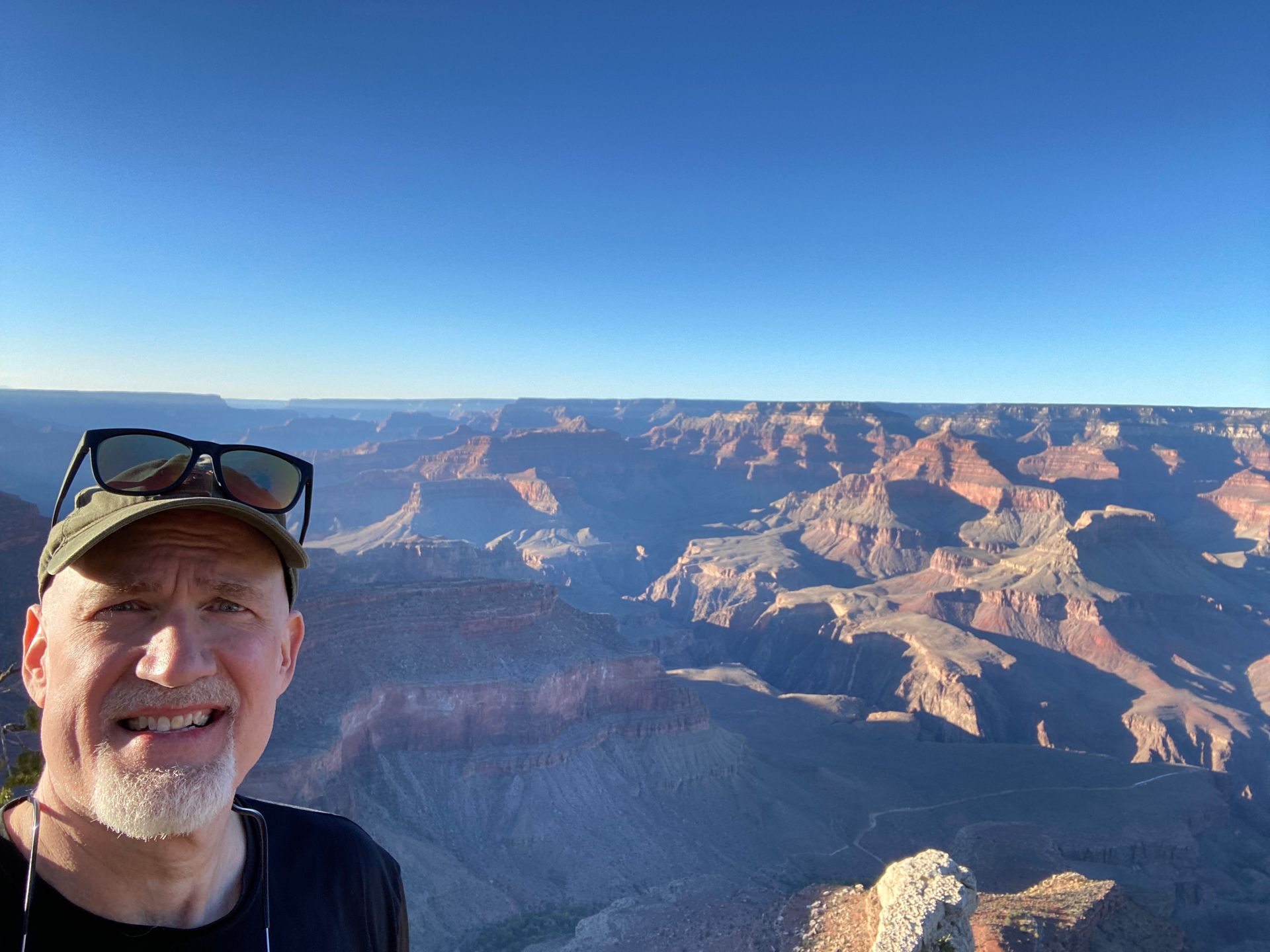 Man with sunglasses smiles at the Grand Canyon under a clear, blue sky.