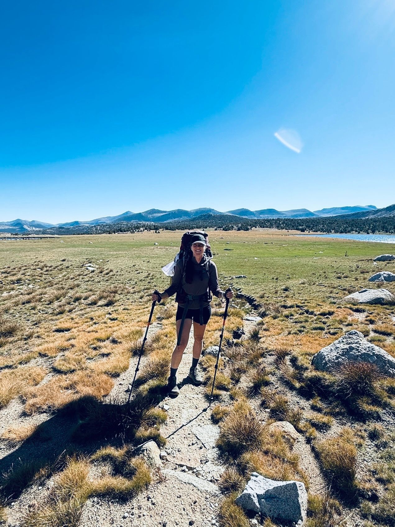 Woman backpacking on a trail with a large pack, using trekking poles, sunny landscape with a lake and mountains.