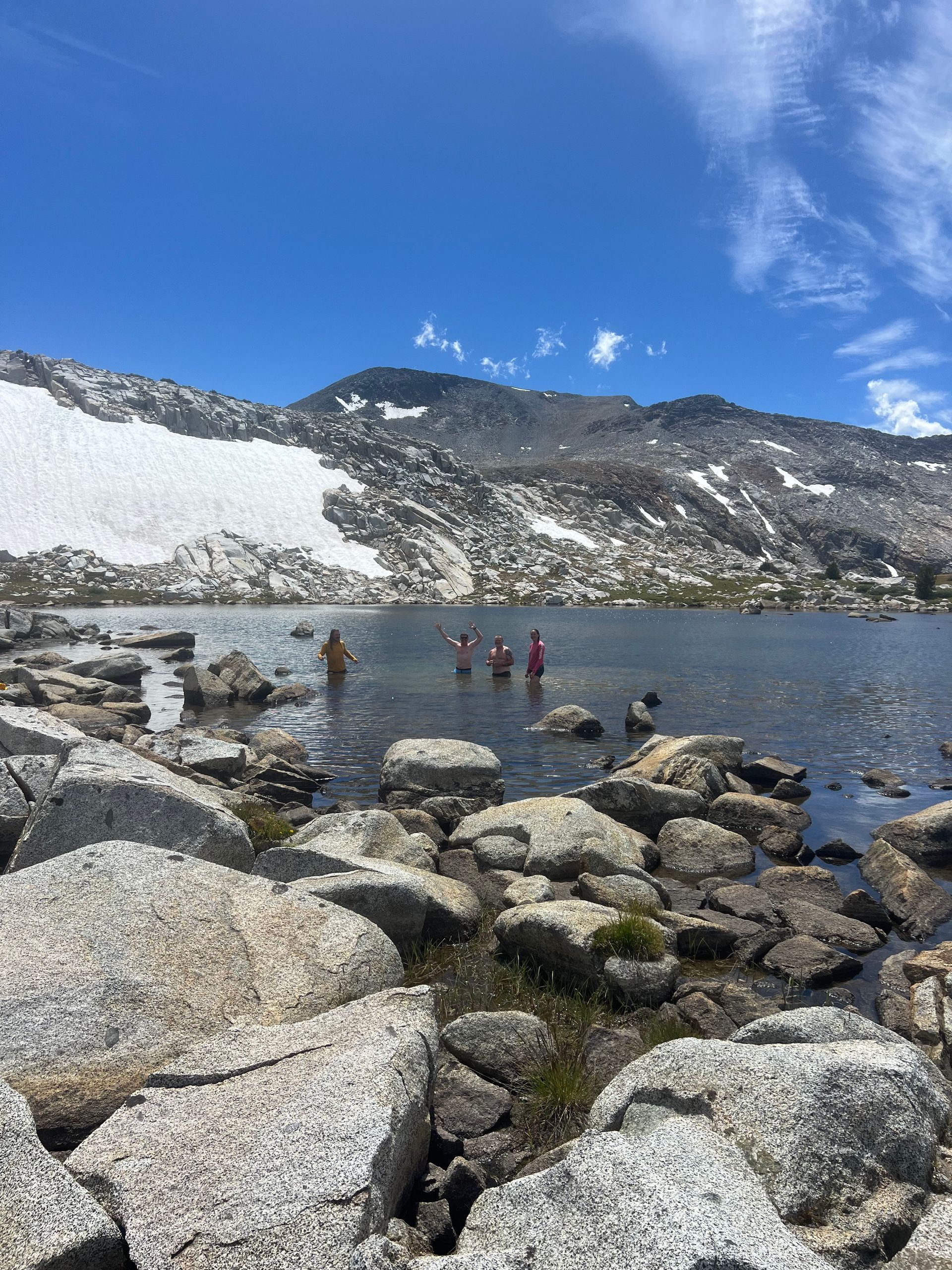 People wading in a clear lake surrounded by large rocks and mountains with patches of snow under a blue sky.