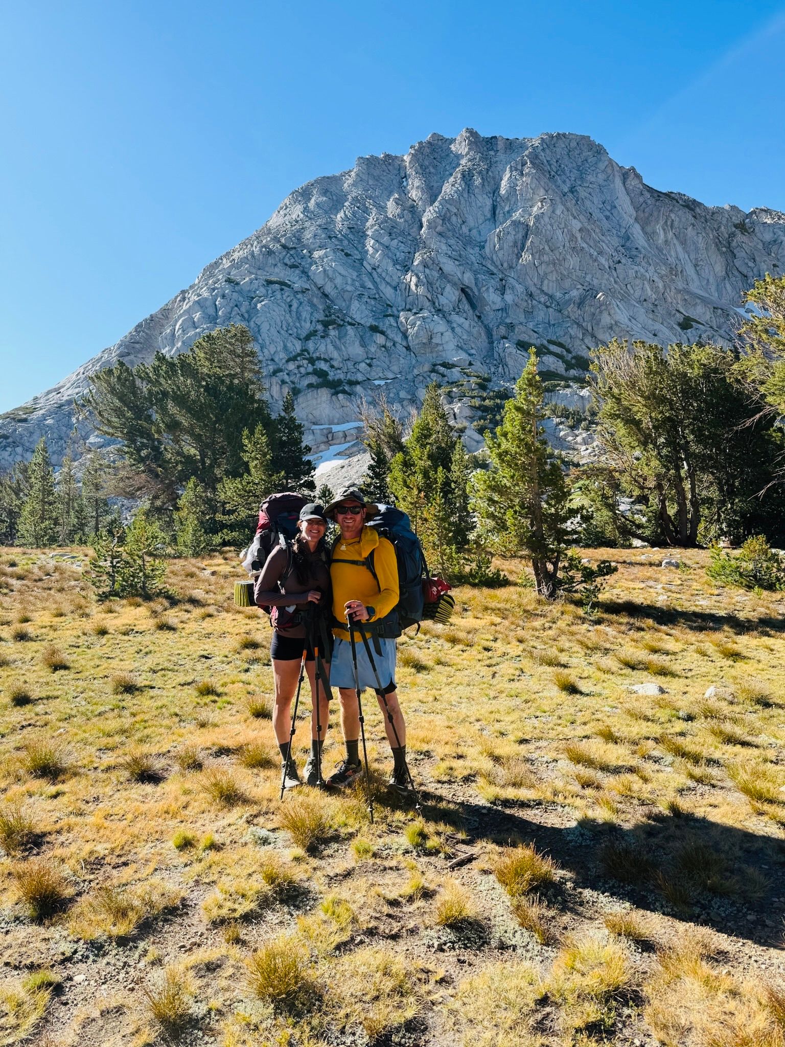 Two hikers with backpacks on a sunny trail, standing before a rocky mountain and pine trees.