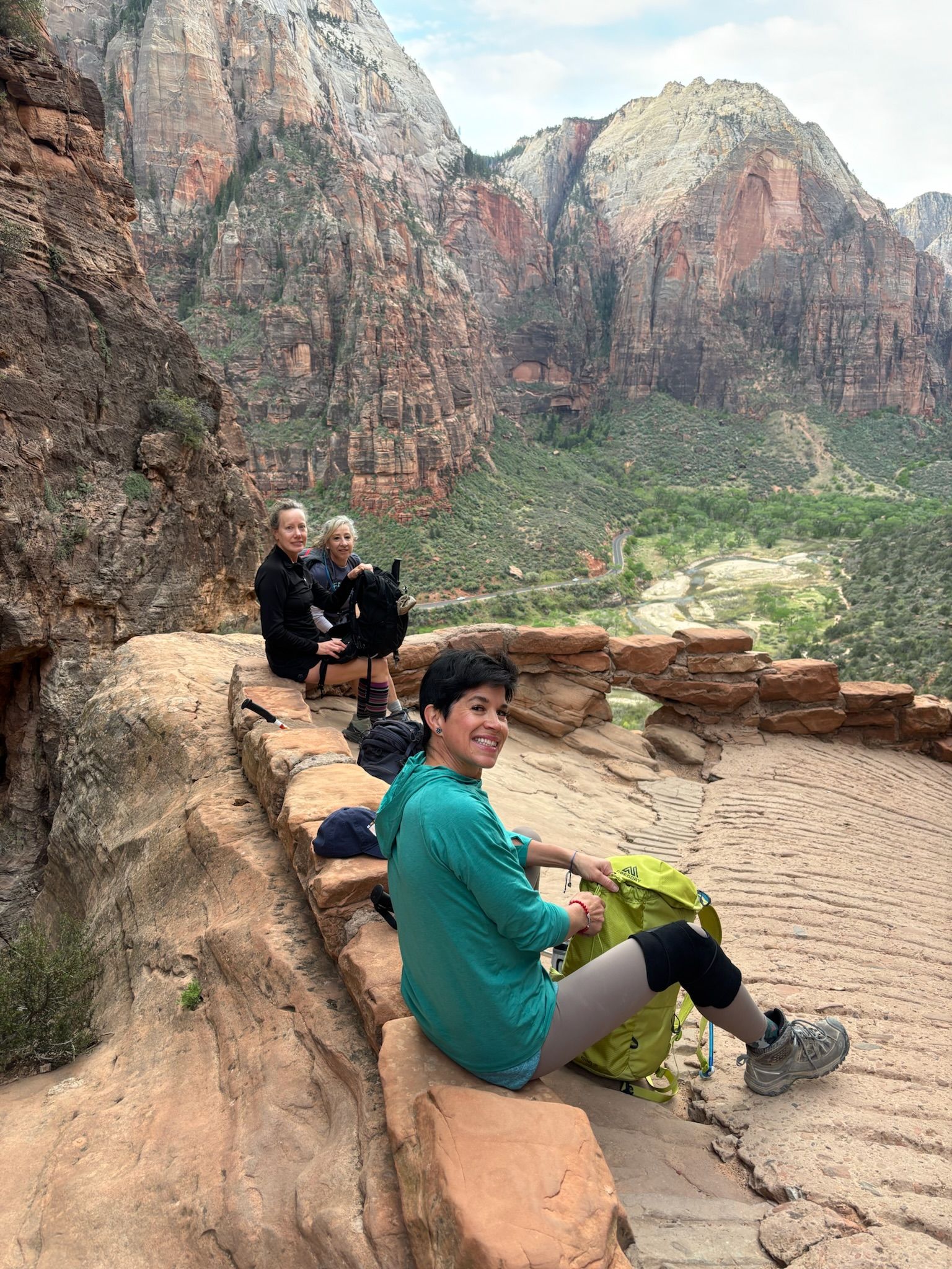Three people rest on a stone ledge overlooking a canyon. One person wears green and gray, smiles at the camera.