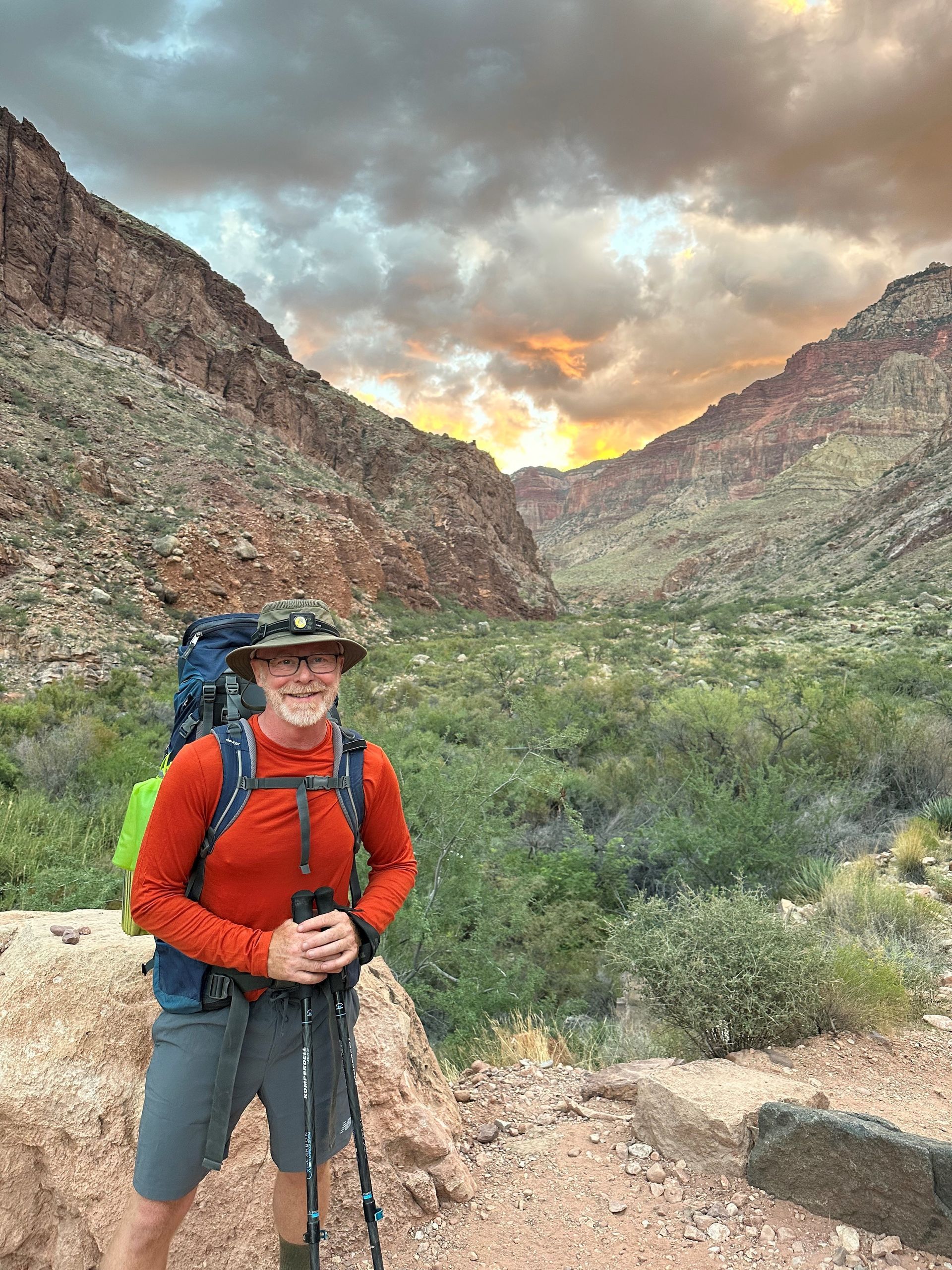 A man with a backpack and hiking poles is standing in front of a mountain.