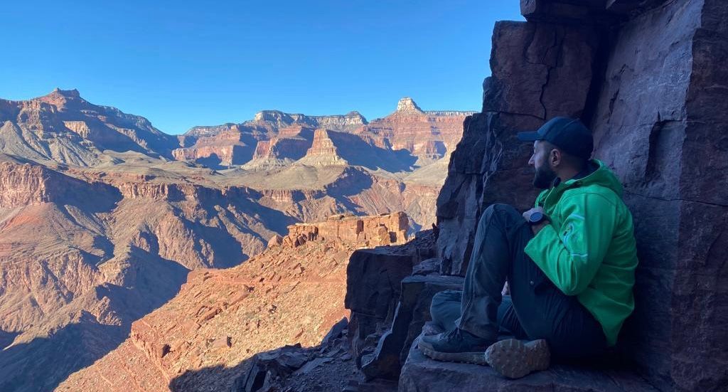 Man in green jacket sits on a ledge, overlooking the Grand Canyon at sunrise.