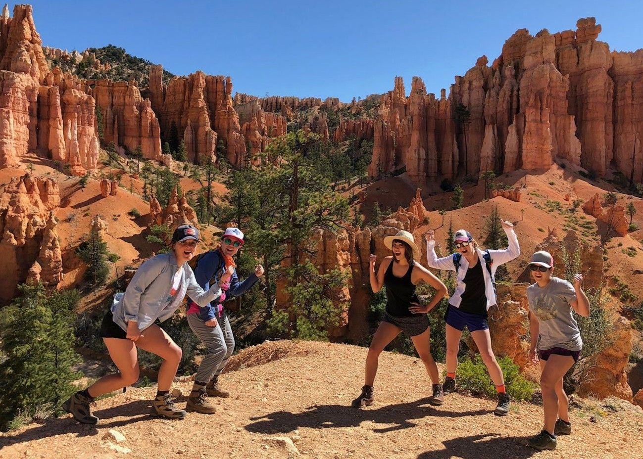 Group posing and dancing in a sunlit red-rock canyon with orange cliffs and blue sky