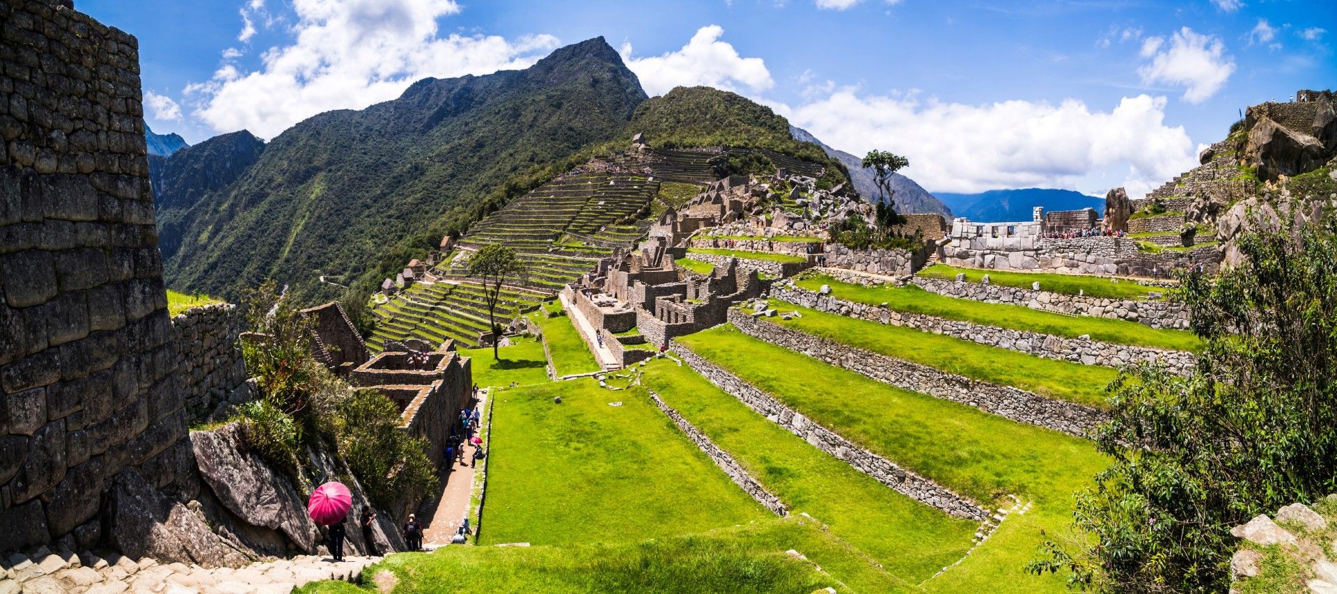 Terraced green hills and stone ruins in a mountain village under a blue sky, with a few hikers.