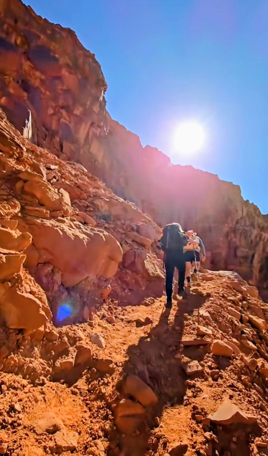 Hikers with backpacks ascend a narrow, sunlit rocky trail between red cliffs under a bright blue sky.