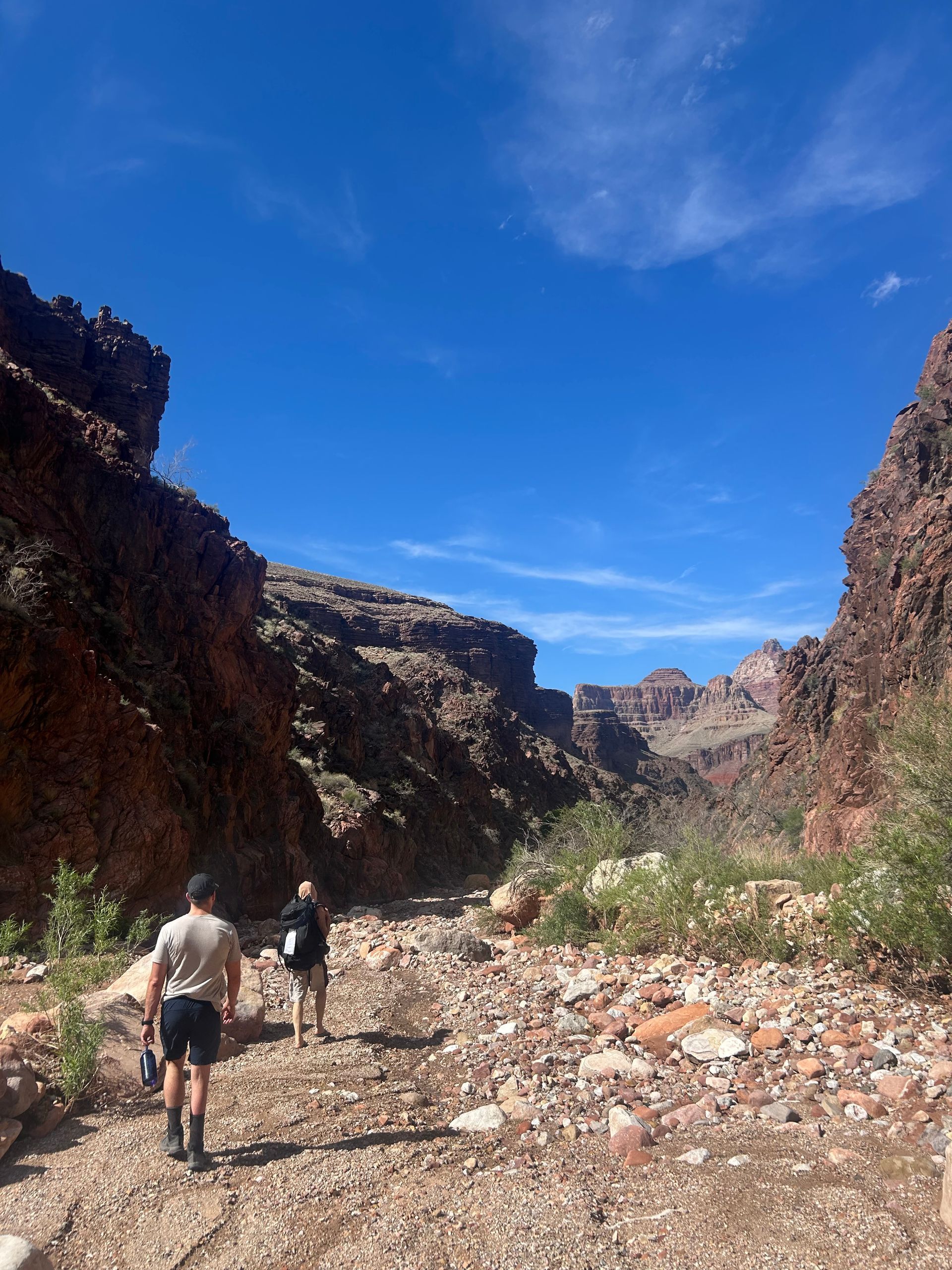Hikers walk along a rocky canyon trail under a bright blue sky.