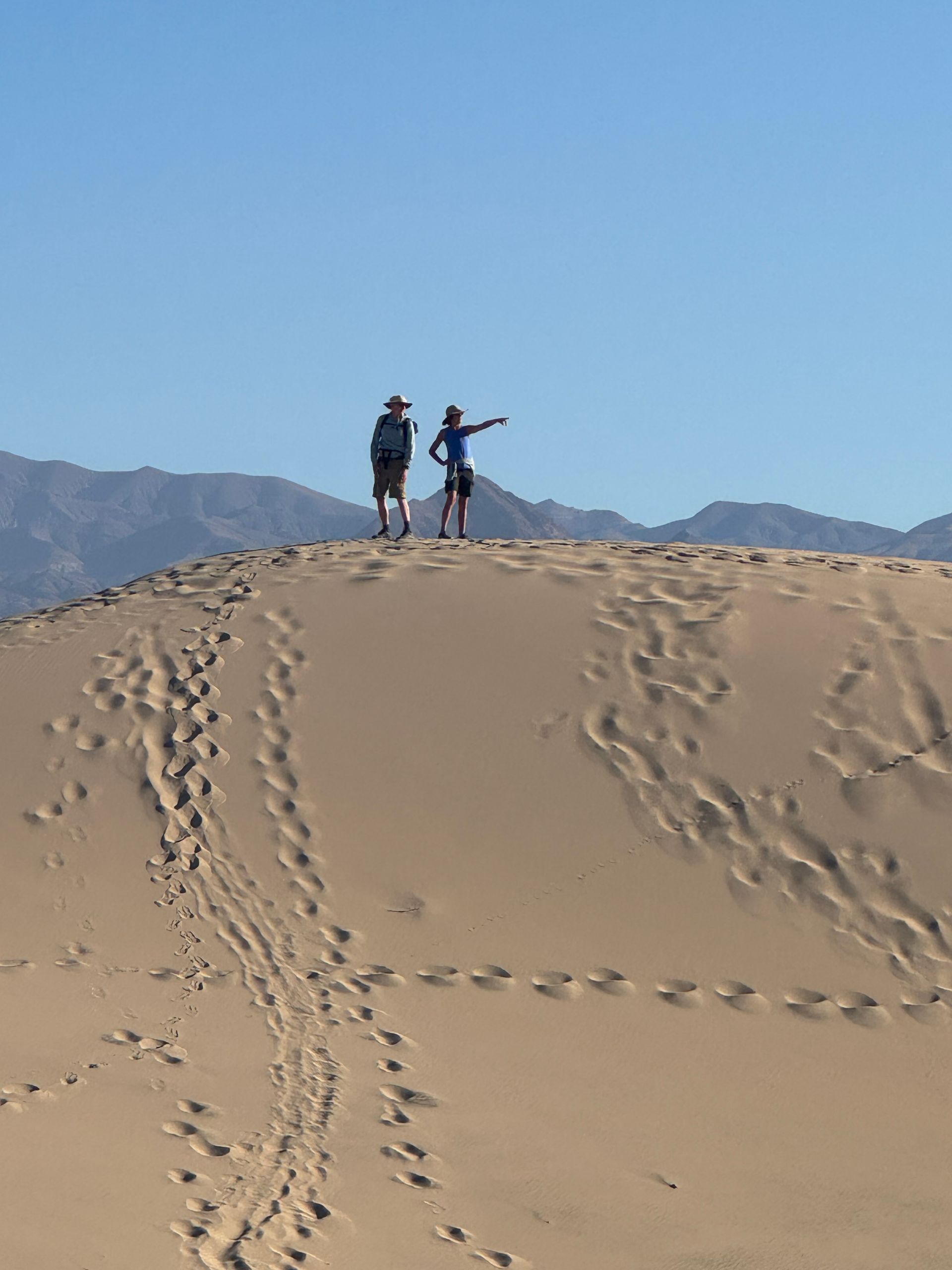 Two people on a sand dune, one pointing. Mountains and a blue sky in the background.