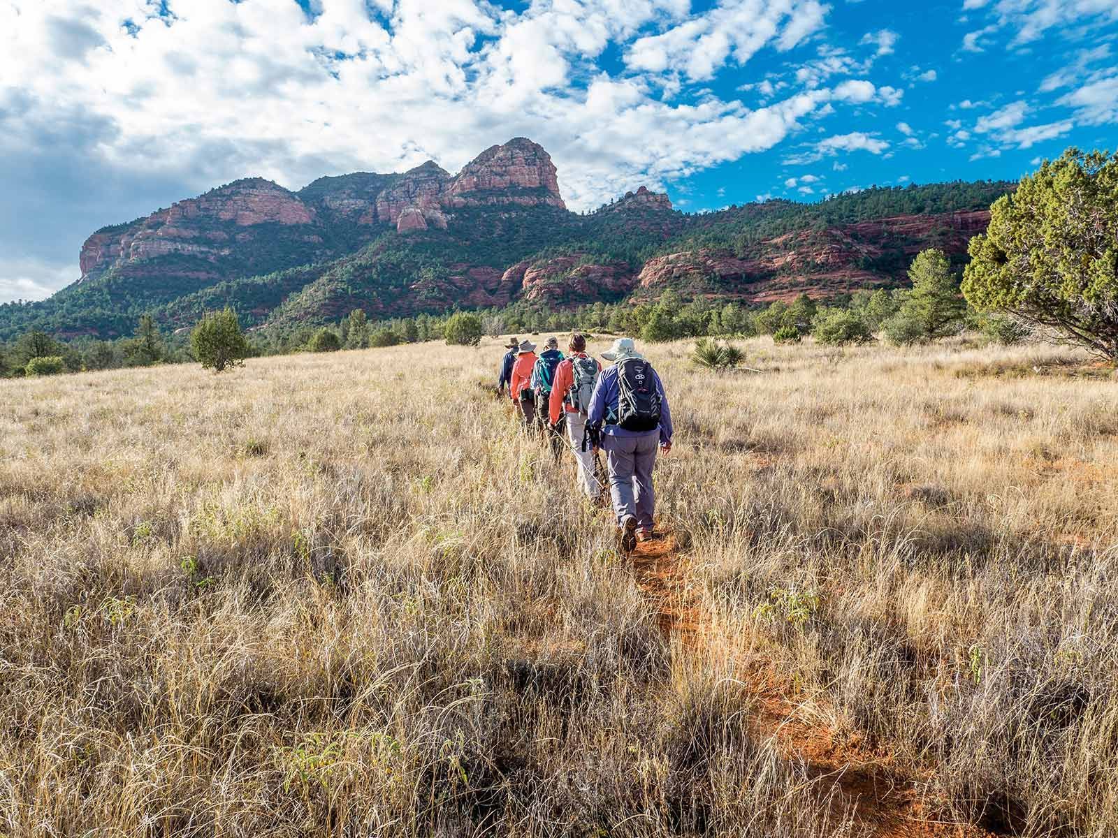 Hikers walking a grassy trail toward red rock mountains under a blue sky