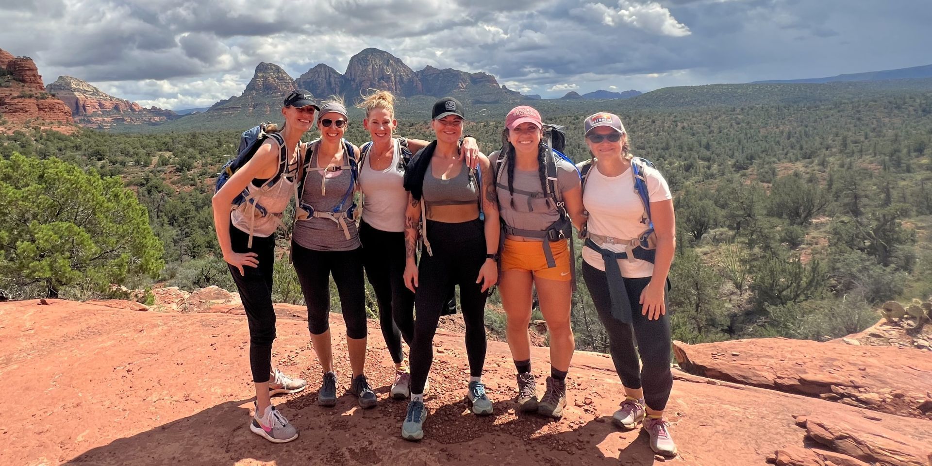 Group of hikers posing on a red rock overlook with canyon cliffs and cloudy mountains behind them