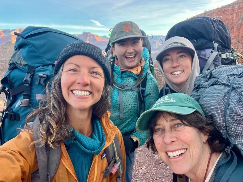 Four hikers smiling for a selfie with backpacks, wearing caps, outdoors in the mountains at dusk