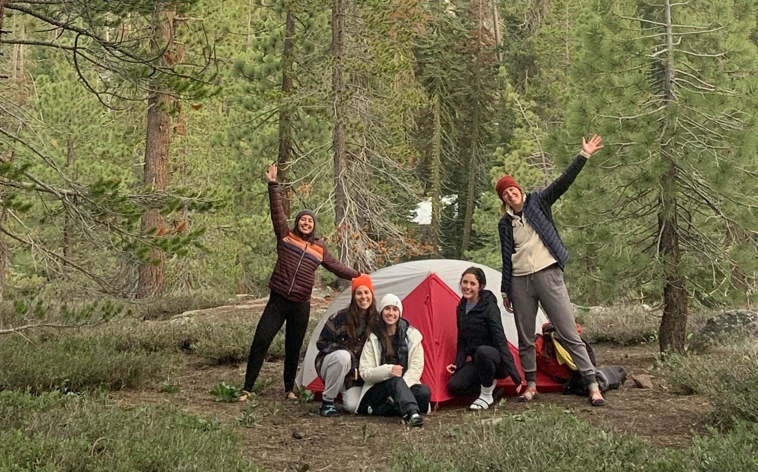 Four people posing by a red tent in a forest, smiling and raising their arms