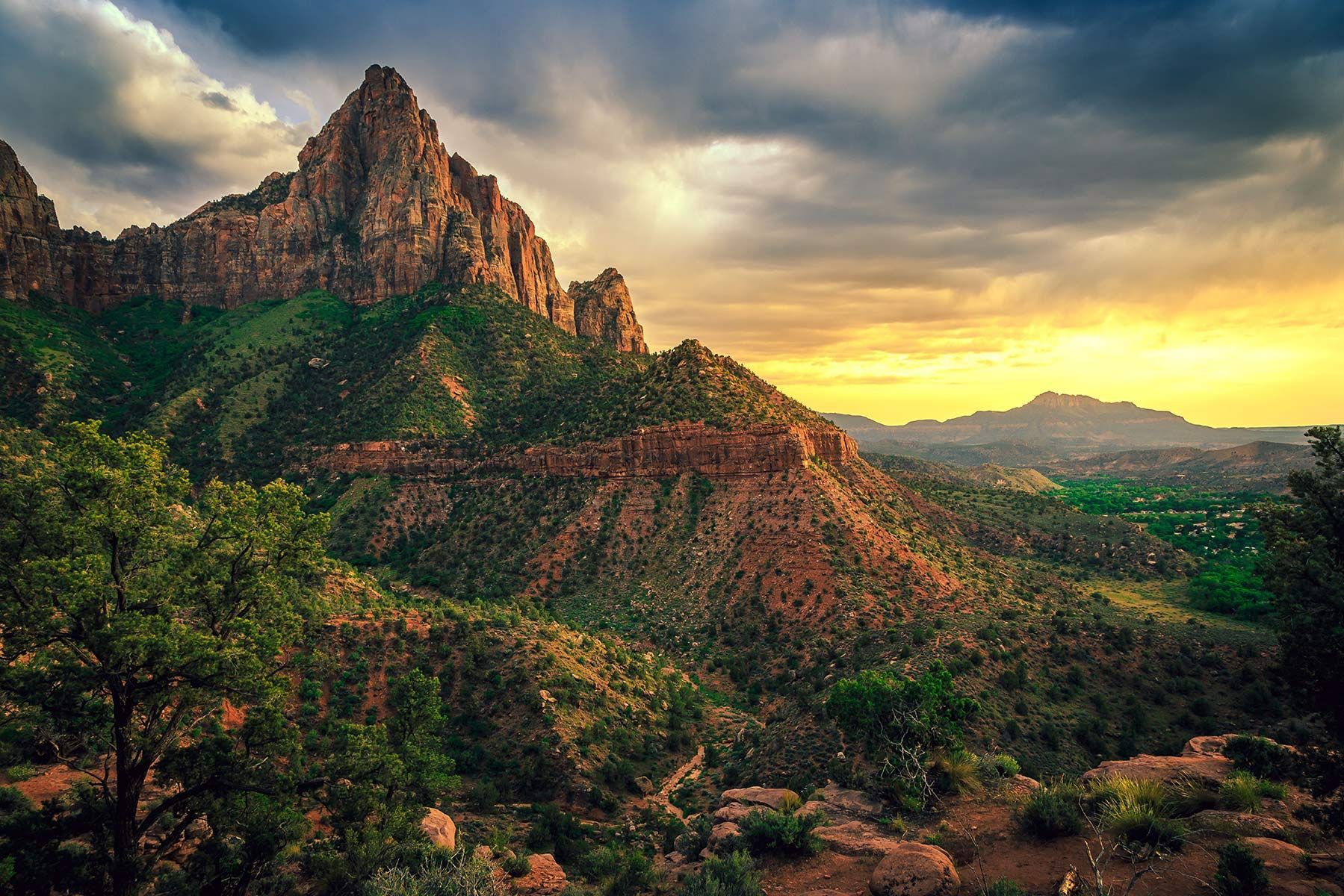 A striking, jagged mountain peak towers over a lush, tree-covered valley at sunset, lit by golden light and dark clouds.