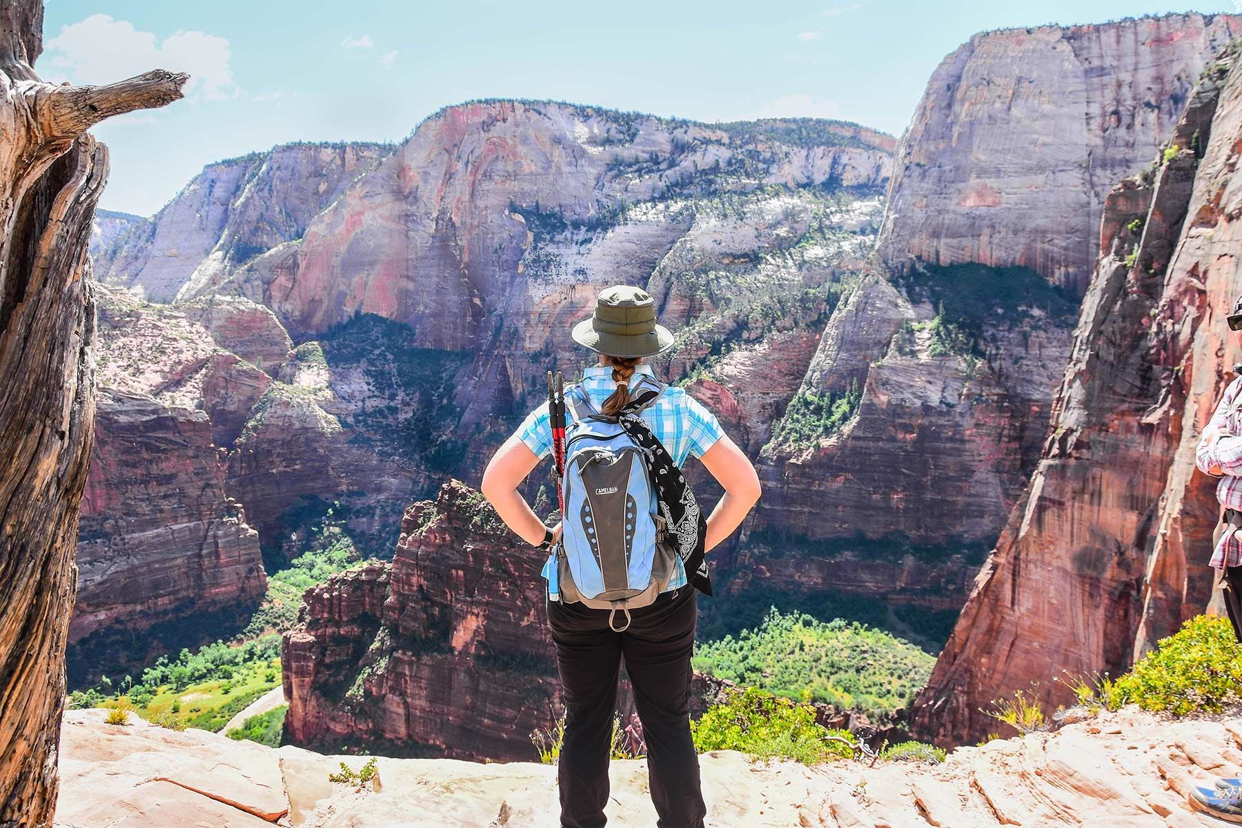 A person with a backpack and hat stands on a rocky ledge, looking out over a vast, sunlit canyon with steep, red cliffs.