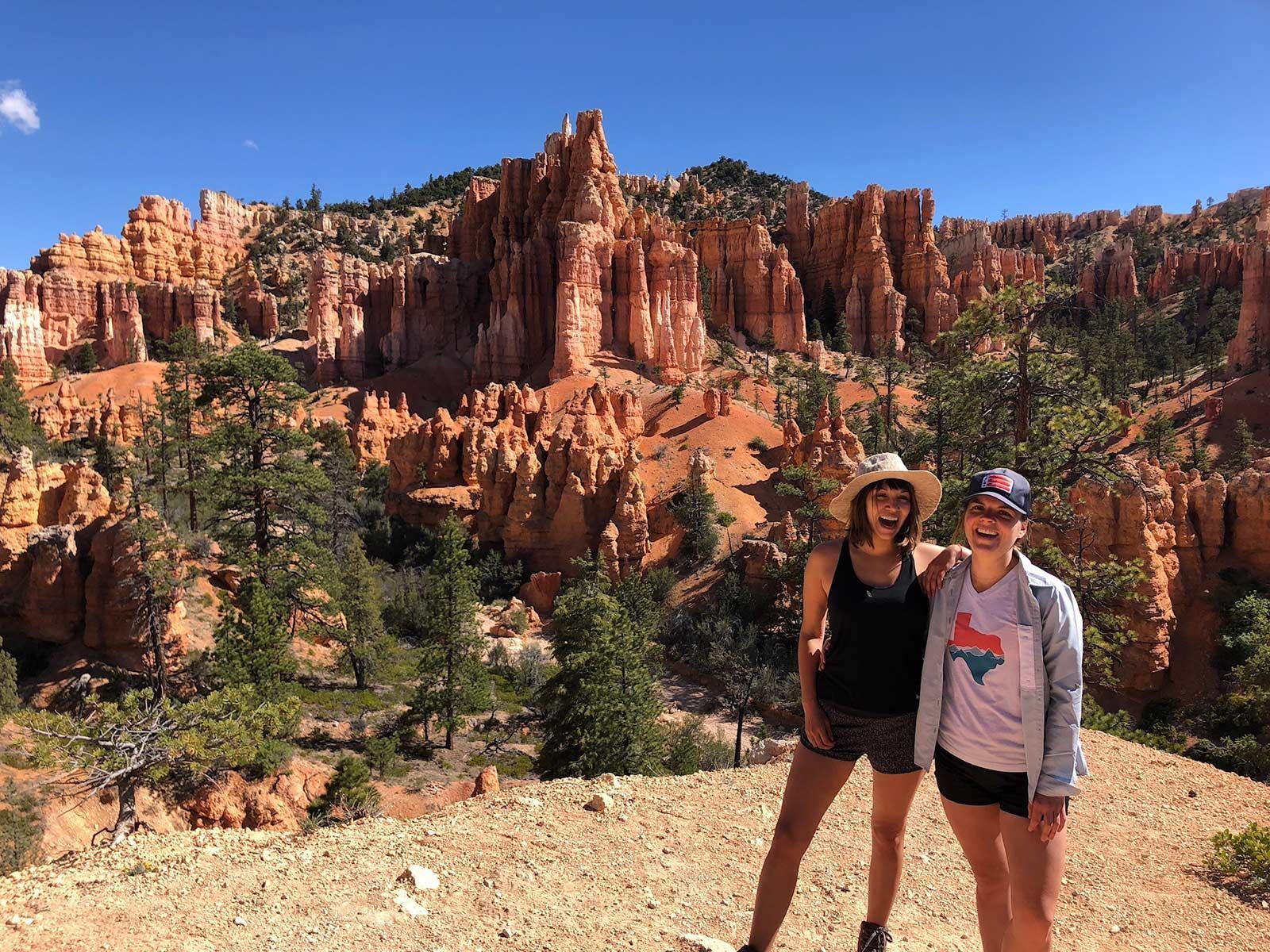 Two women smile posing in front of red rock formations and trees, under a bright blue sky.