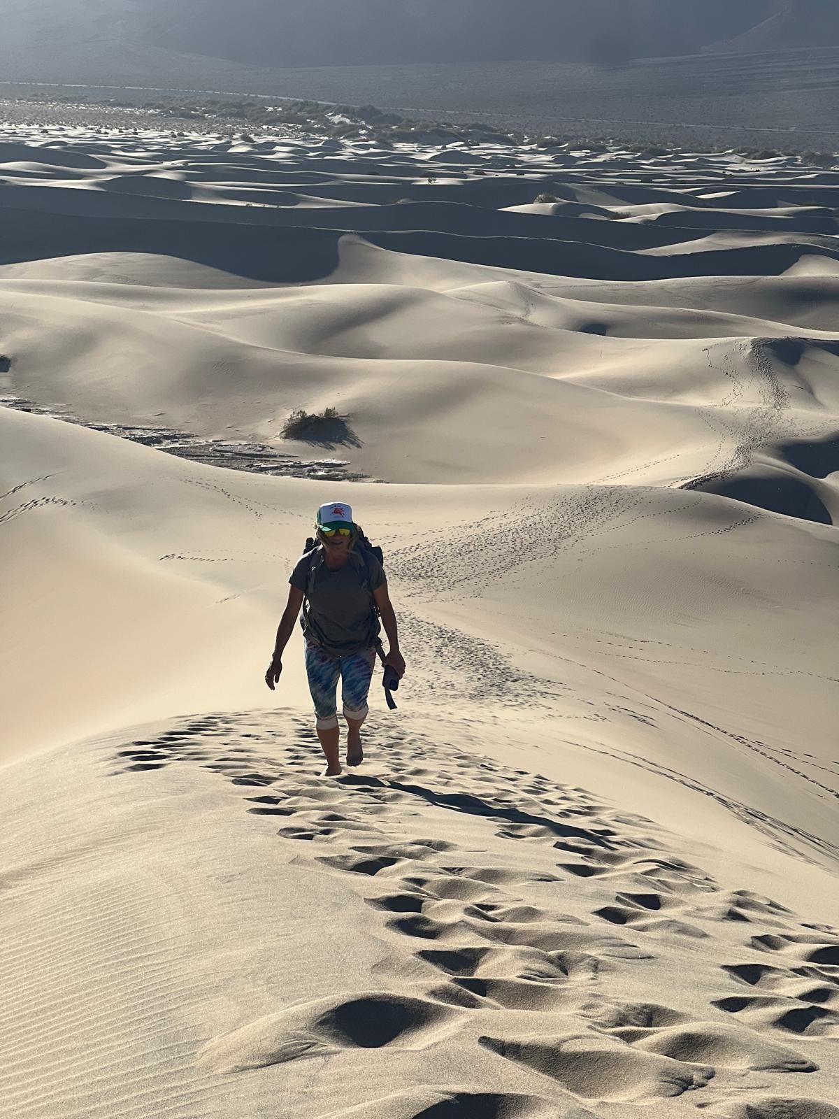 Hiker descending a sandy dune ridge under a cloudy sky, with footprints trailing behind.
