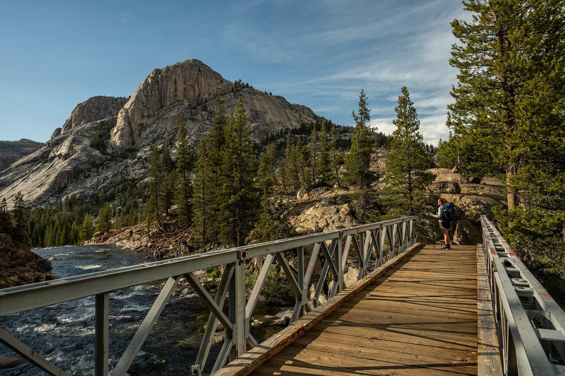 Wooden bridge beside a river in a mountain forest, with a person walking toward rocky cliffs.