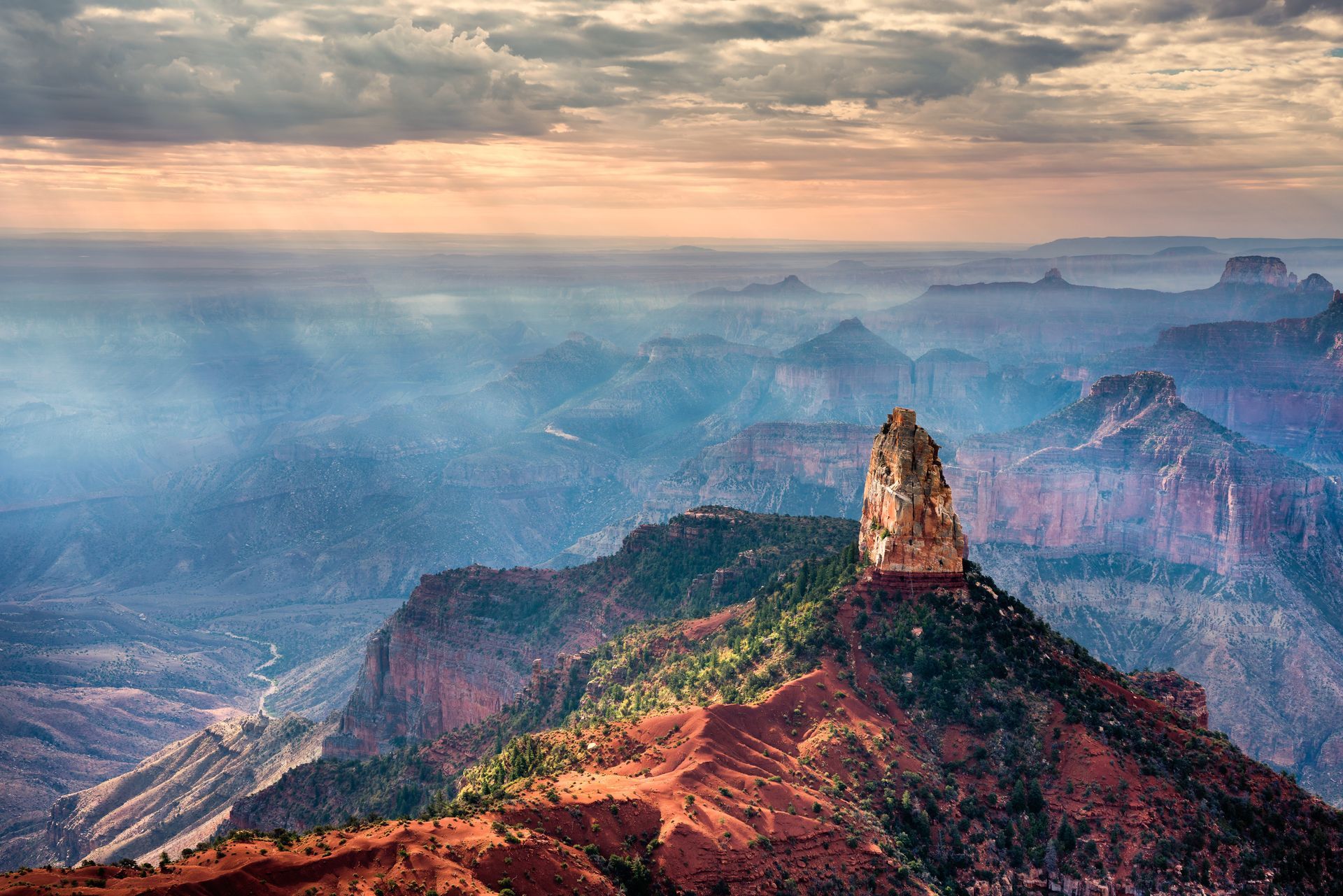 Canyon landscape with a tall, prominent rock formation in the foreground, under a cloudy sky.