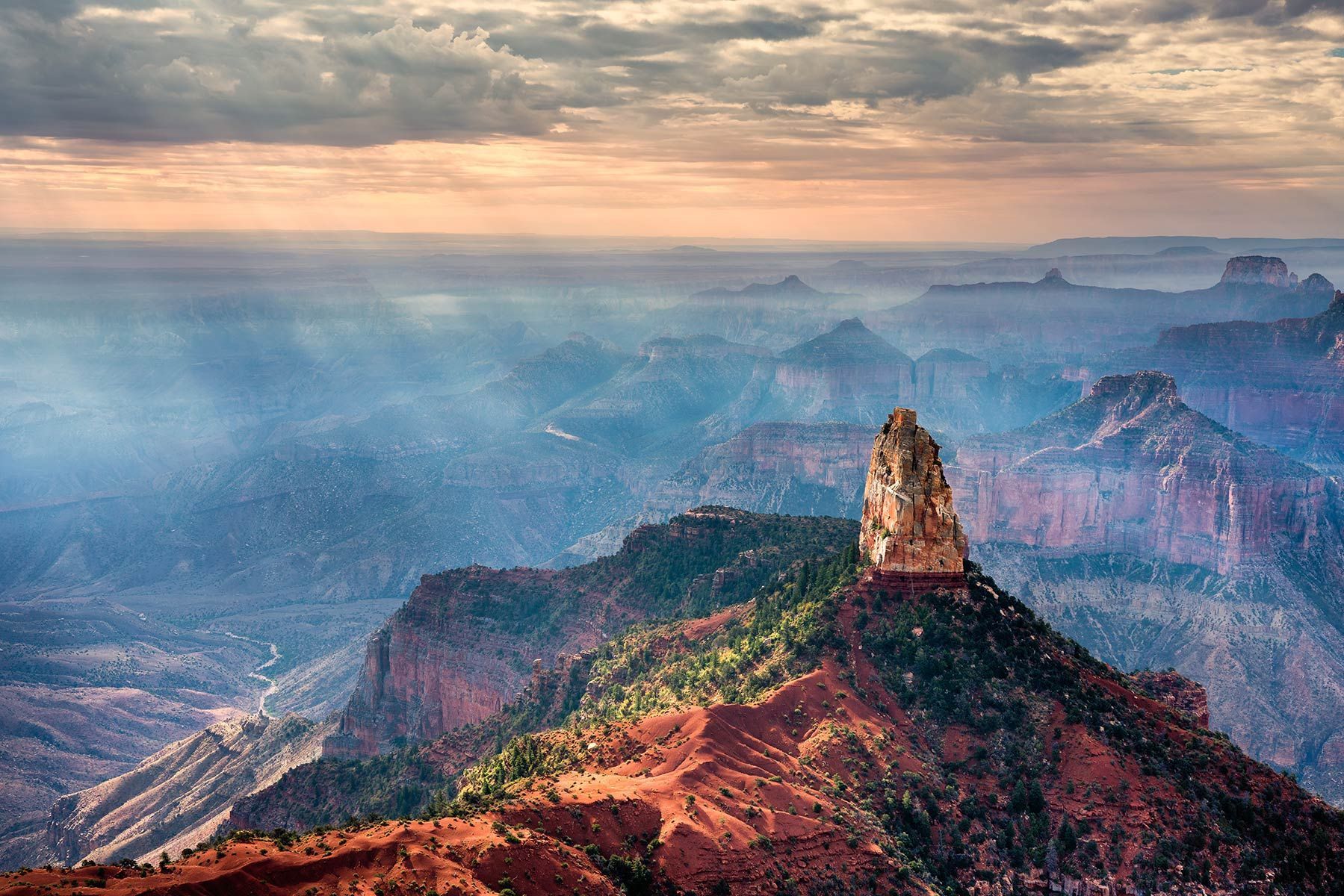 Sunlit desert canyon landscape with a tall rock spire and hazy blue mountains at sunrise