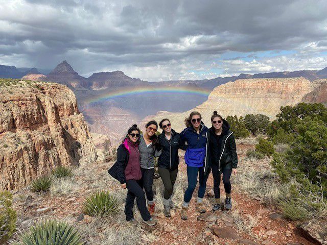 Group of hikers posing on a rocky overlook with a rainbow and canyon cliffs under cloudy skies
