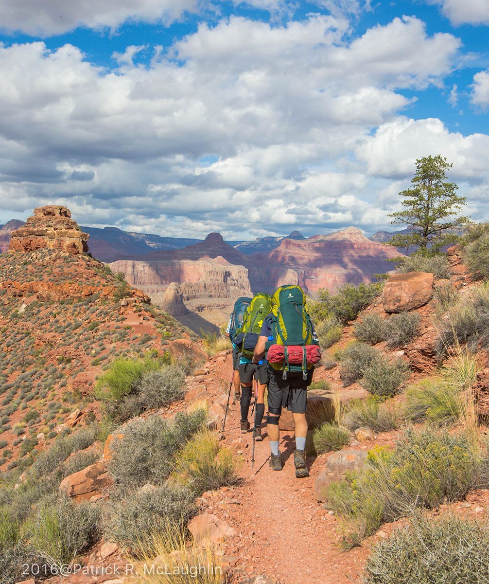 Hikers with backpacks on a trail, red rock landscape, sunny with clouds.