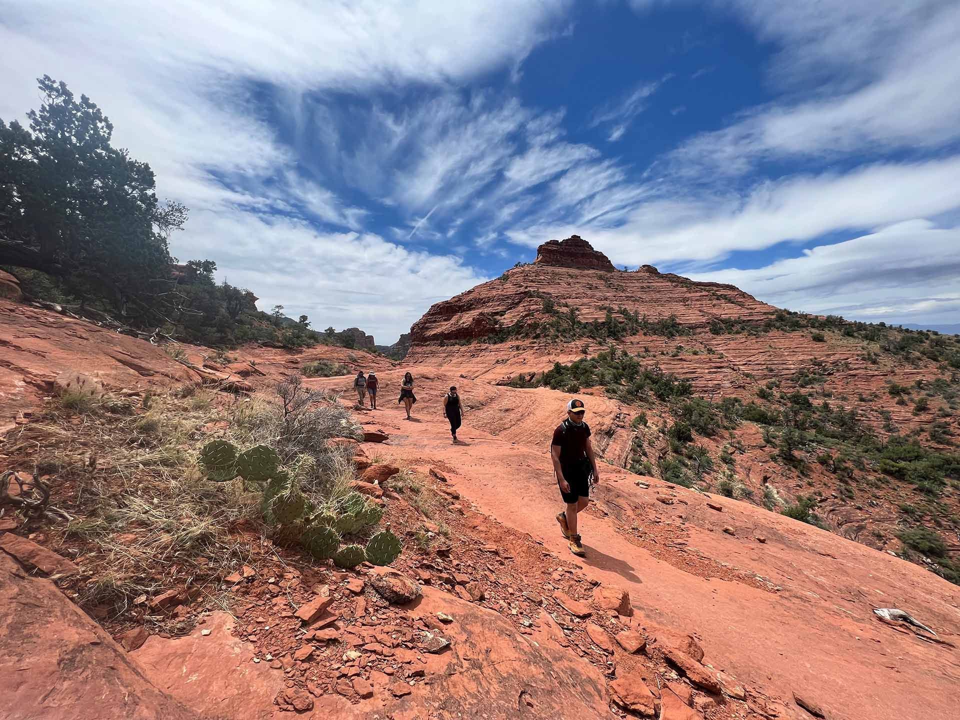 Hikers walking along a red sandstone trail beneath a blue, cloud-streaked sky